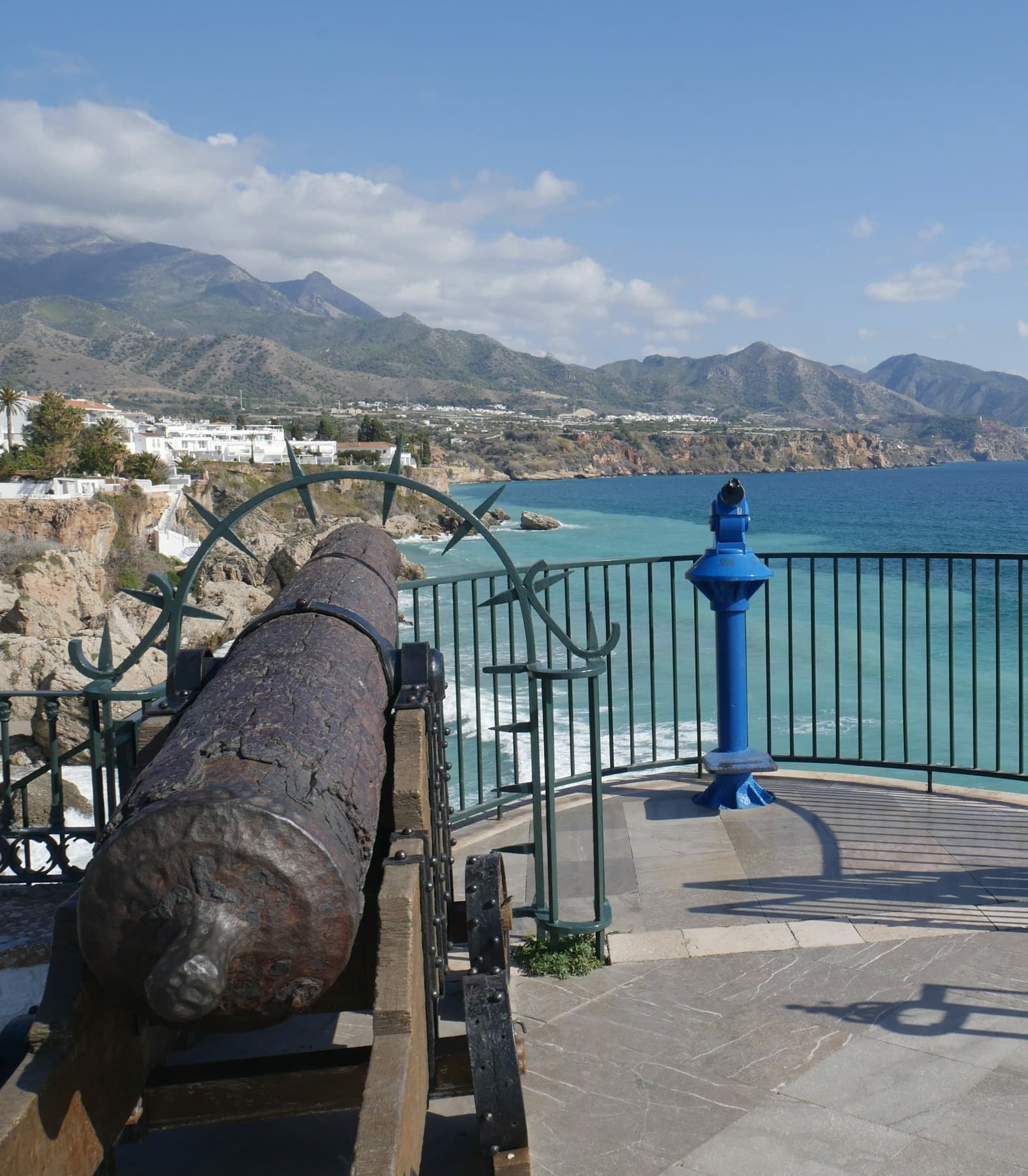 This image depicts a viewing area with a antique canyon, telescope and view of the coastline, mountains and ocean on the Balcony of Europe (Balcón de Europa) in Nerja.