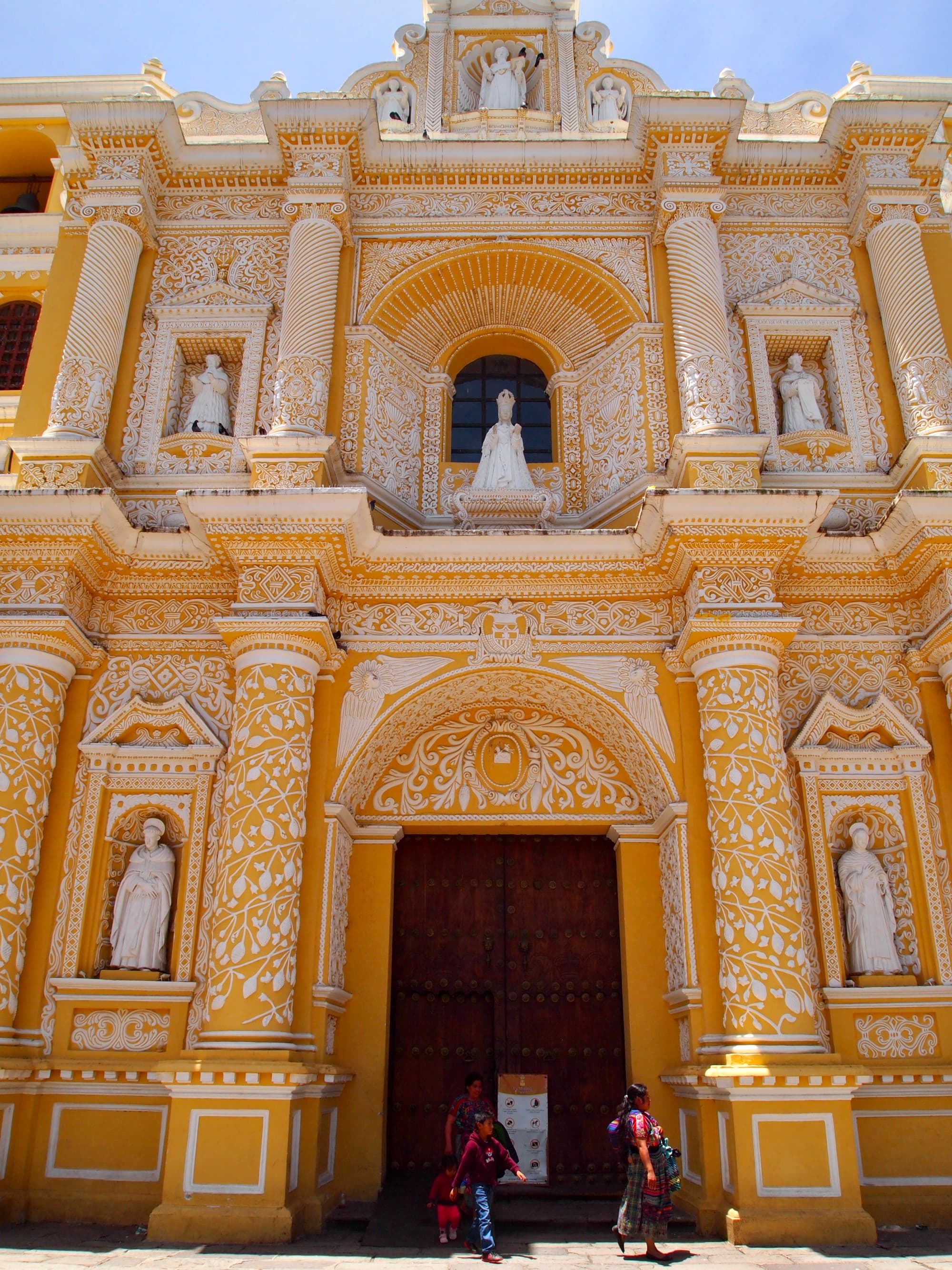 The image features an ornate yellow church facade with intricate designs and statues, in front of which stand two individuals.