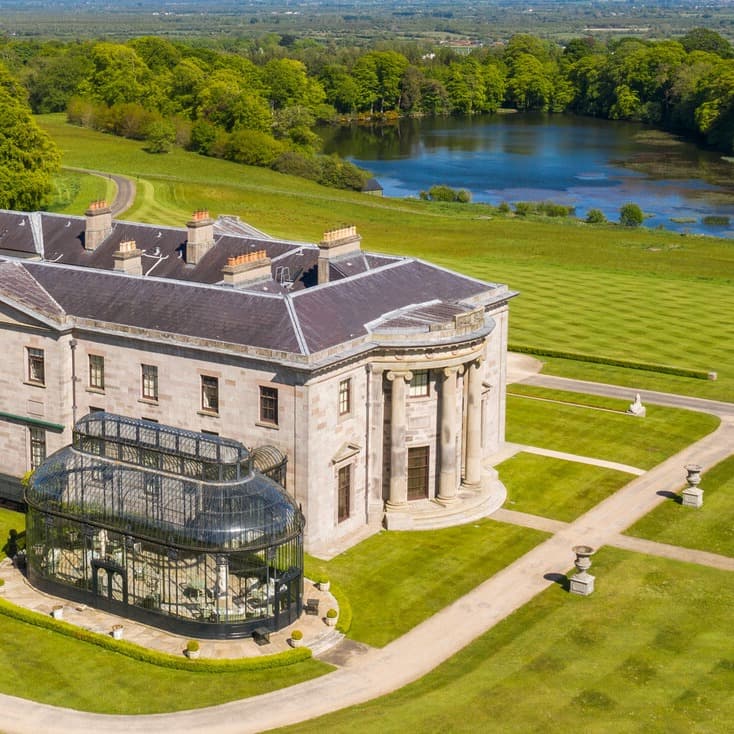 A large manor on a green lawn with a lake in the distance