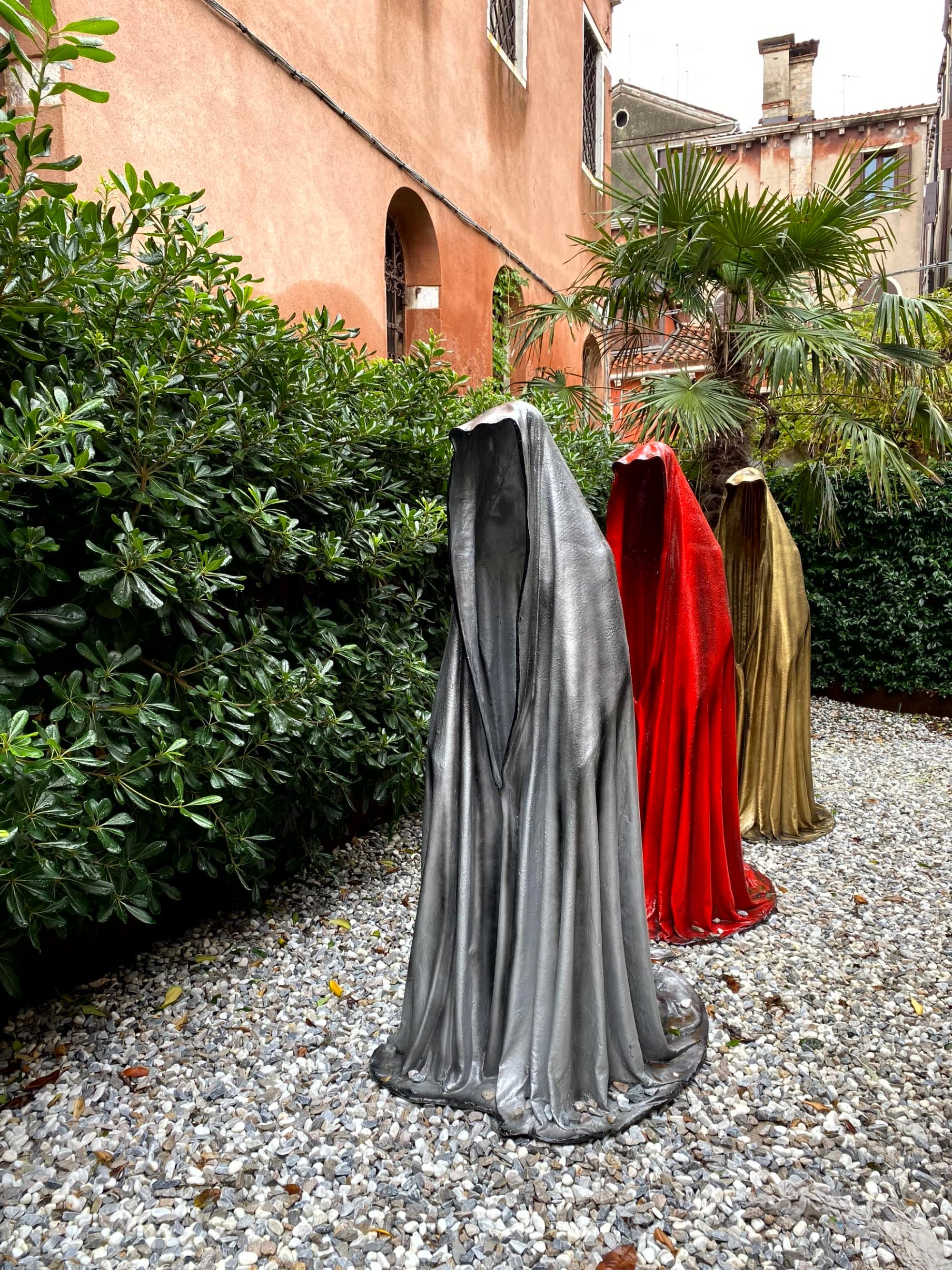 A sculpture of three headless, armless robed figures set against a backdrop of greenery and an orange-walled building.