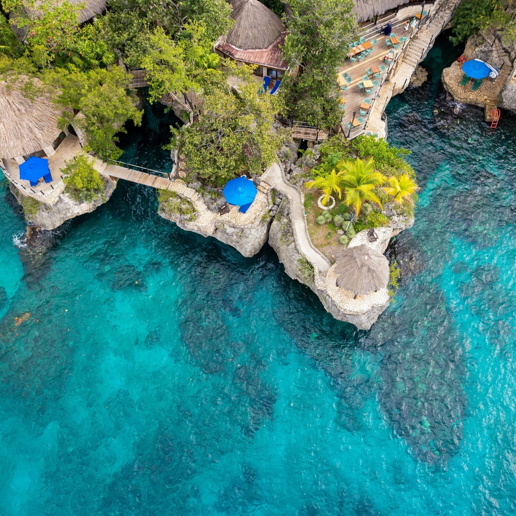 The image is an aerial view of a coastal resort with thatched-roof structures and blue umbrellas, surrounded by clear turquoise waters.