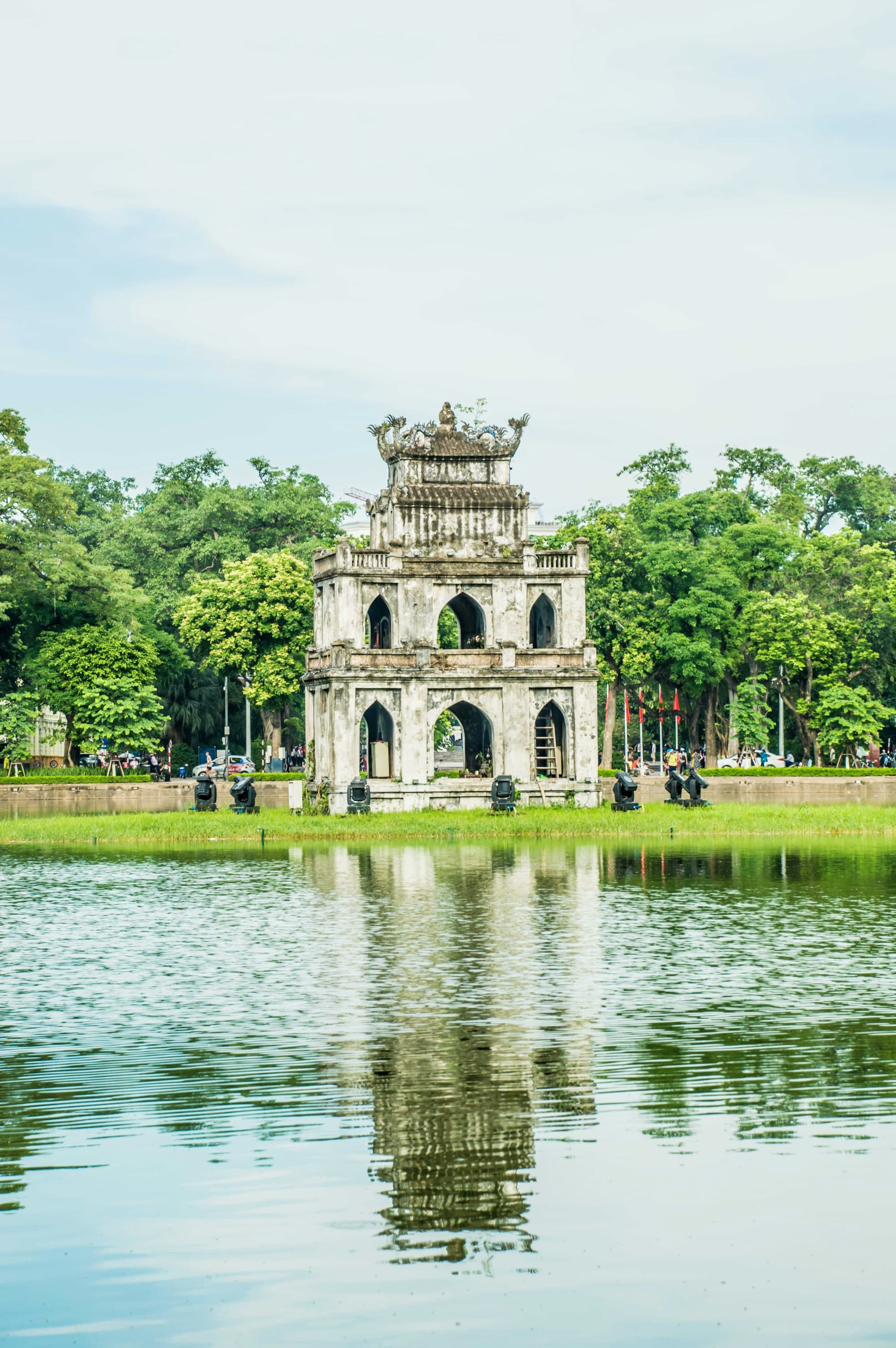 A three-tiered, ornate gate-like structure reflected in a body of water, surrounded by greenery and a clear sky.