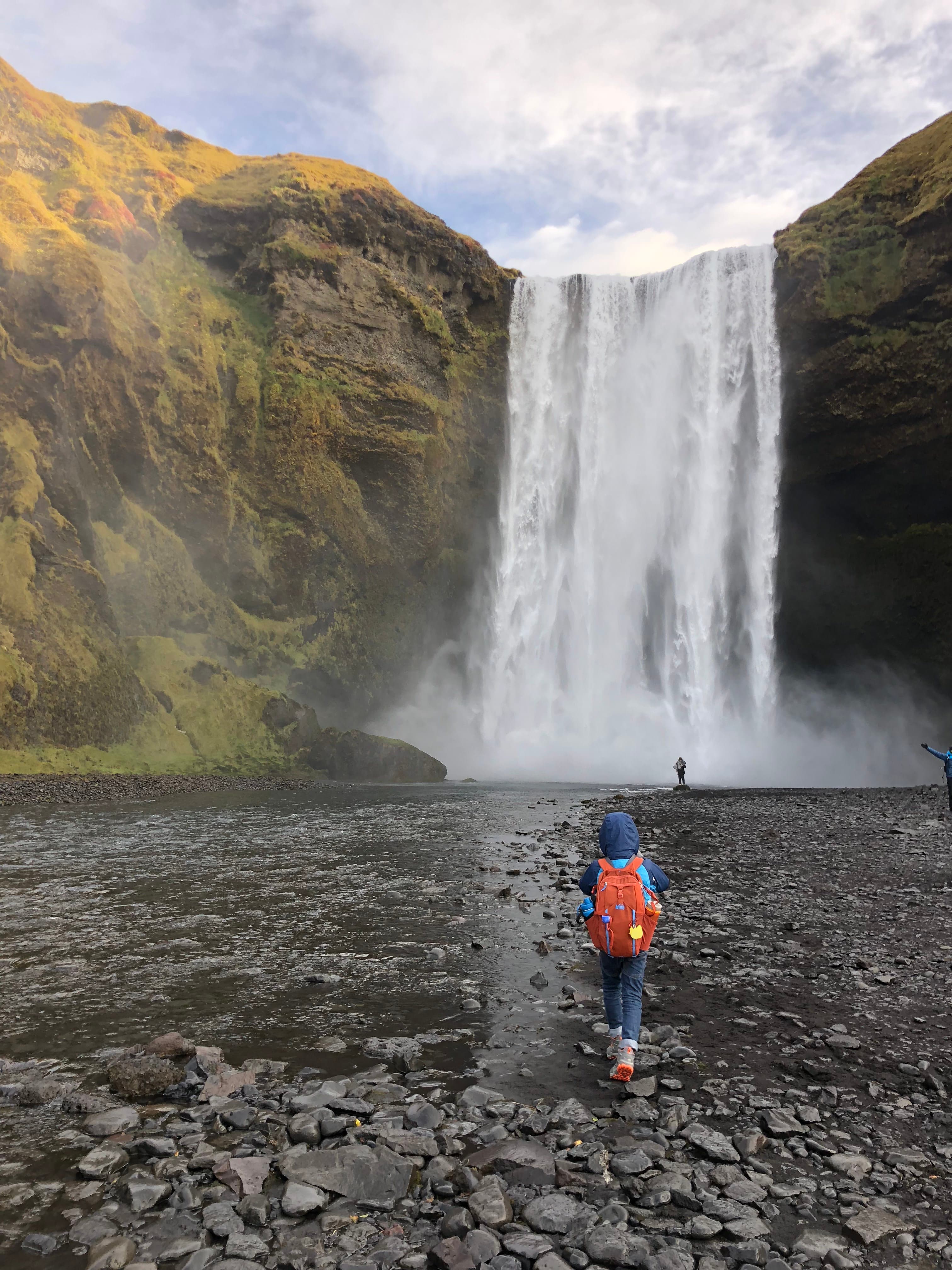 A kid hiking towards Skogafoss Waterfalls on a rocky shore as clouds cover the afternoon sky.