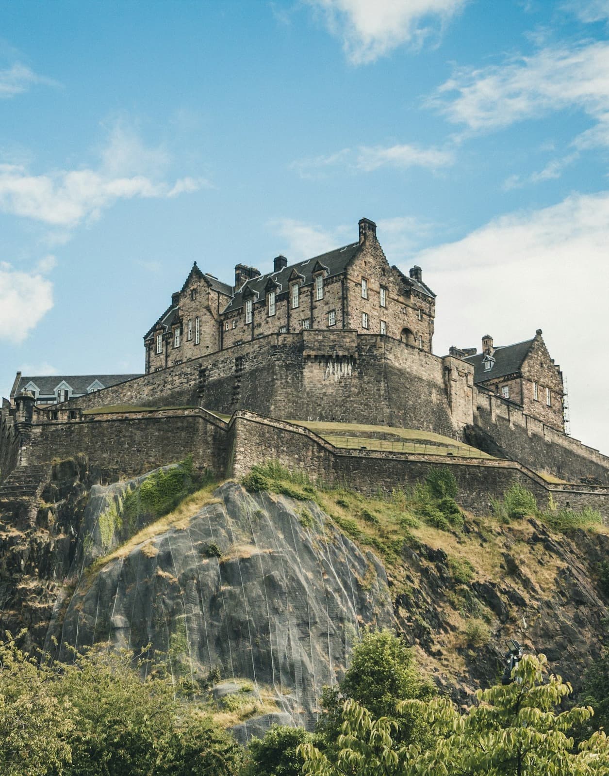 Edinburgh Castle, a historic fortress perched atop Castle Rock in Edinburgh, sits atop a rocky hillside on a sunny day dotted with clouds.