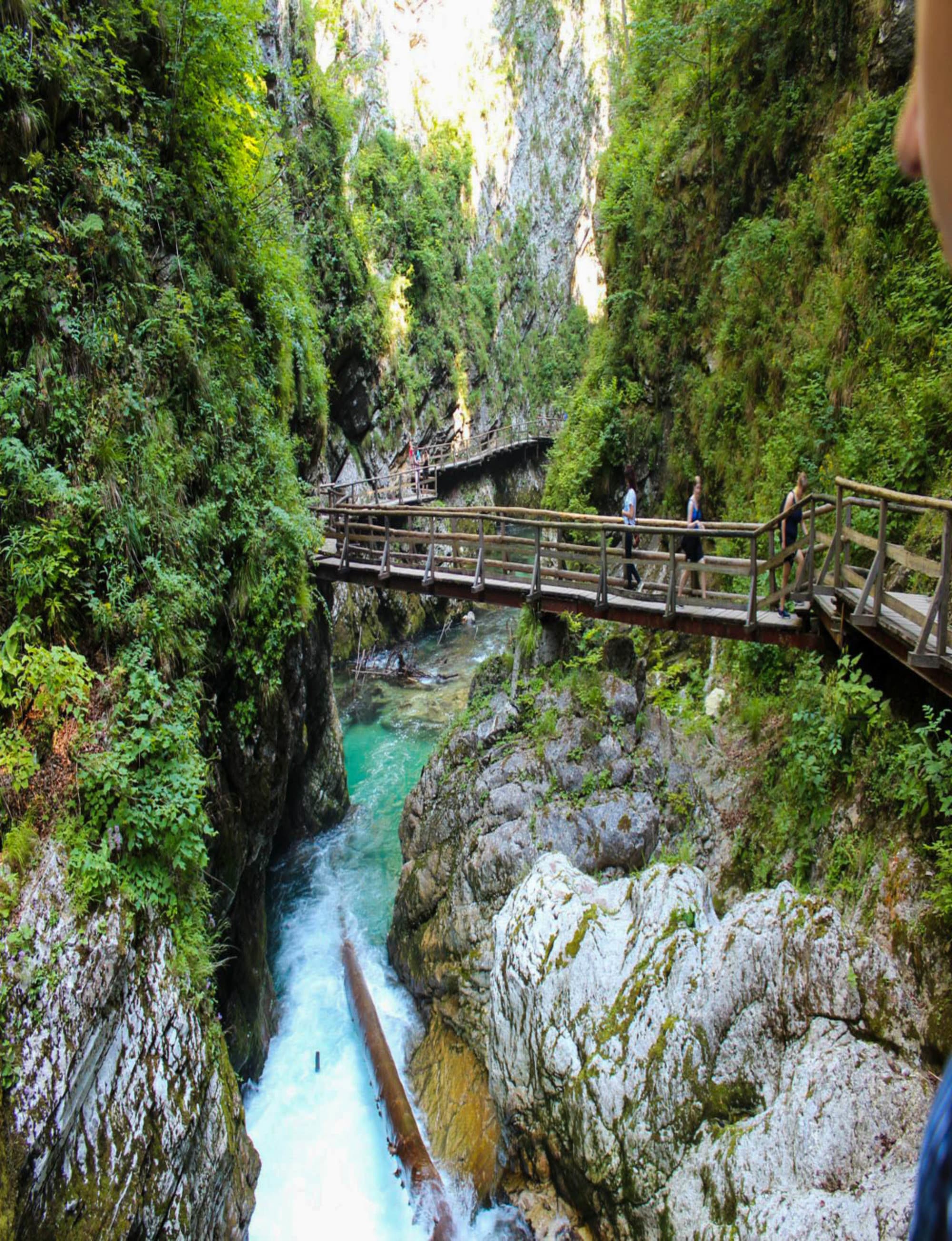 The image features a wooden walkway along a rocky gorge with a turquoise river below, amidst lush greenery and visitors enjoying the view.