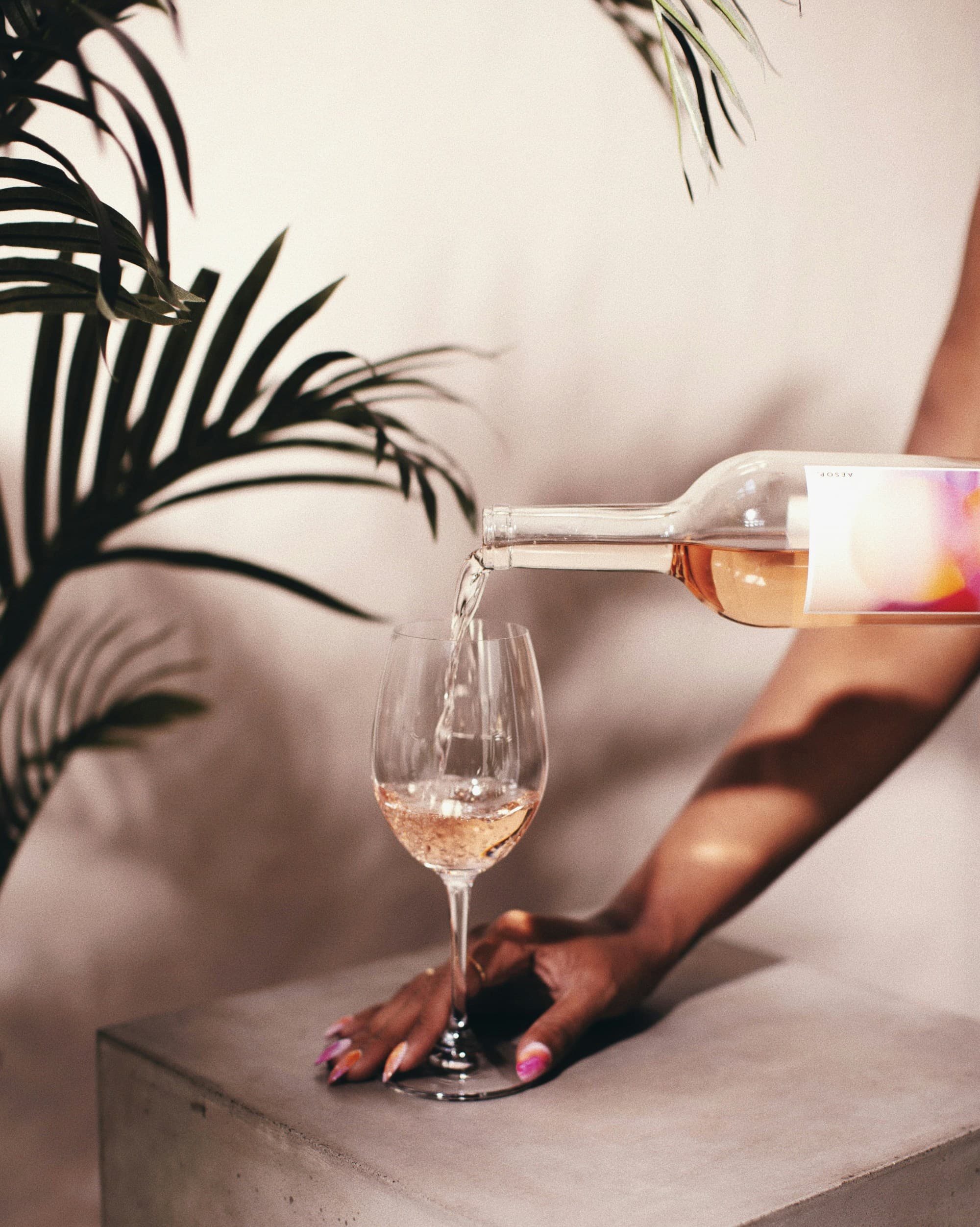 The image captures a moment of someone pouring a clear liquid, likely wine, into a glass against a backdrop of lush foliage.