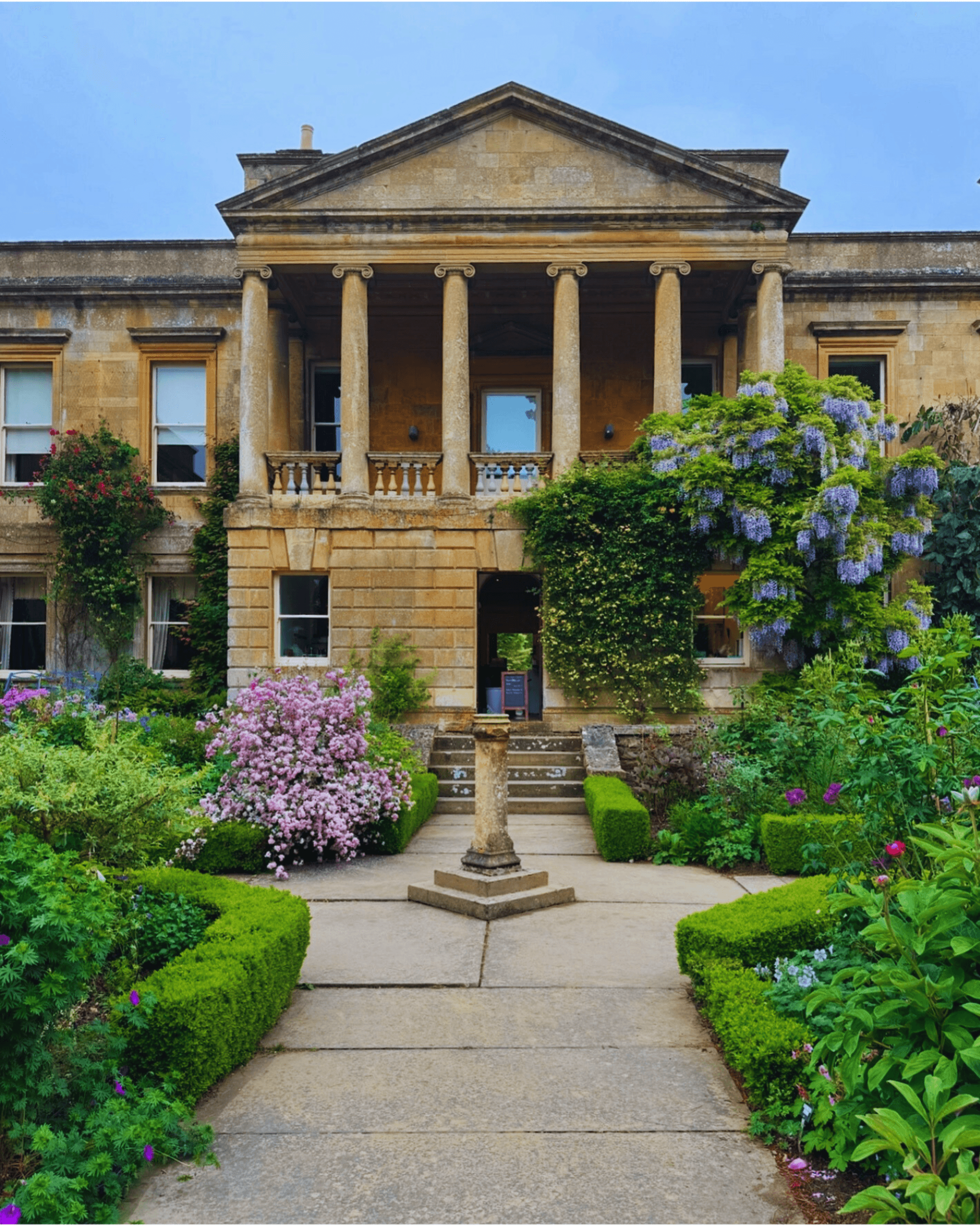 The image depicts the neoclassical Kiftsgate Court Gardens in Gloucestershire, England, surrounded by vibrant floral arrangements.
