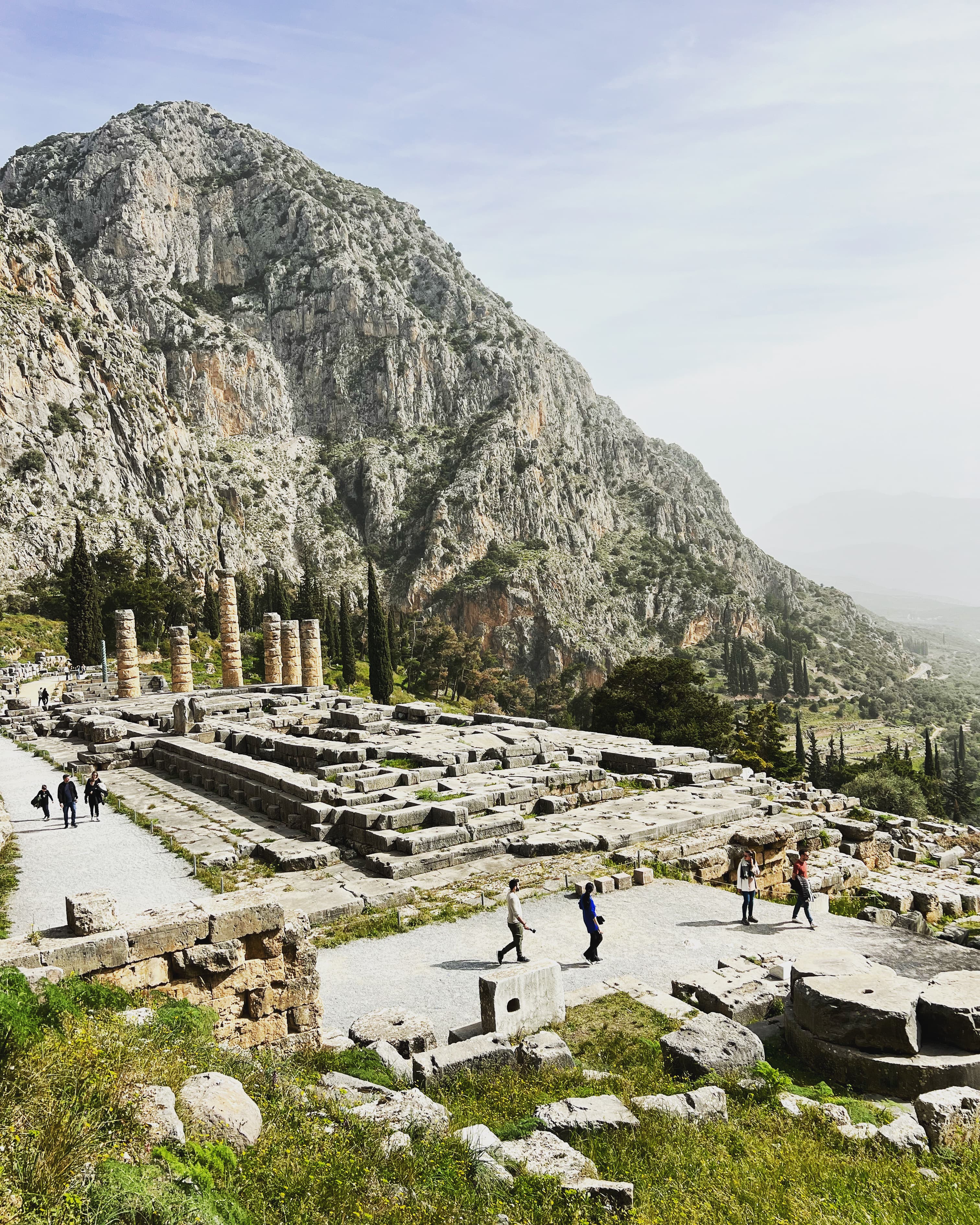 A view of people walking around Ancient Greek ruins outside. There is a mountain in the background covered in scattered trees and brush.