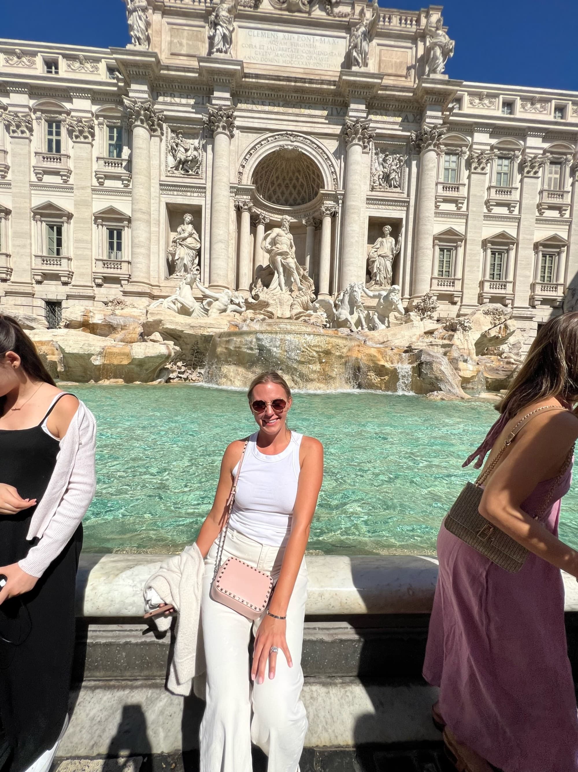 A woman posing in front of the trevi fountains light blue water and white architecture, while wearing a monochromatic white outfit and light pink purse.