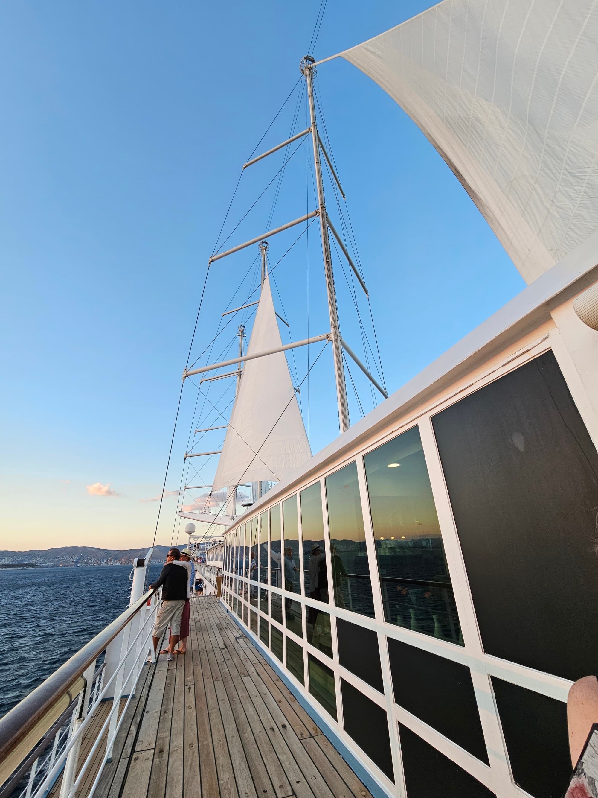 The image depicts a sunset view from the deck of a sailboat with sails unfurled and the horizon in the background.