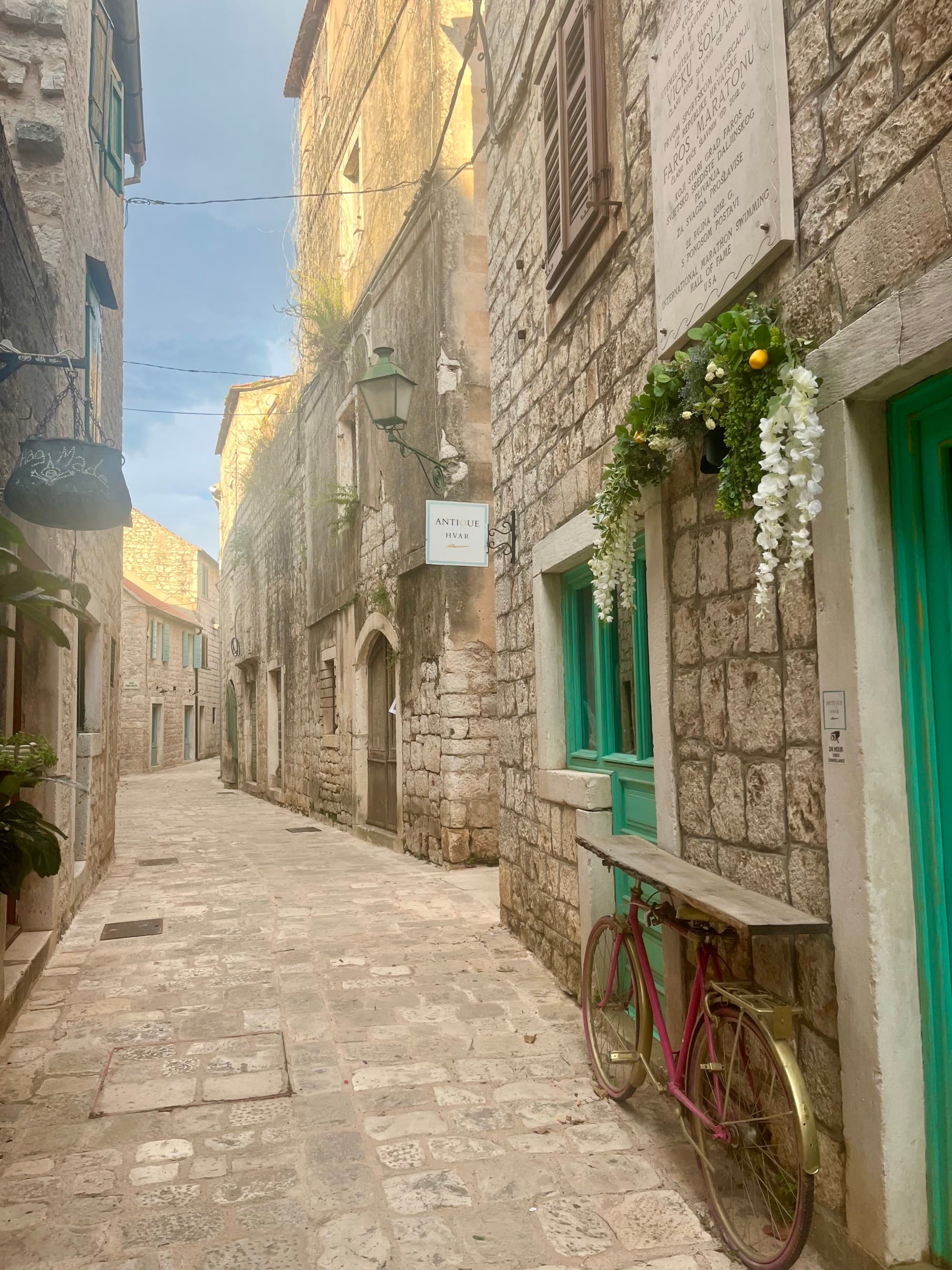 Picturesque stone buildings line a cobbled street with colorful doors and flowers hanging from window boxes on a sunny day.