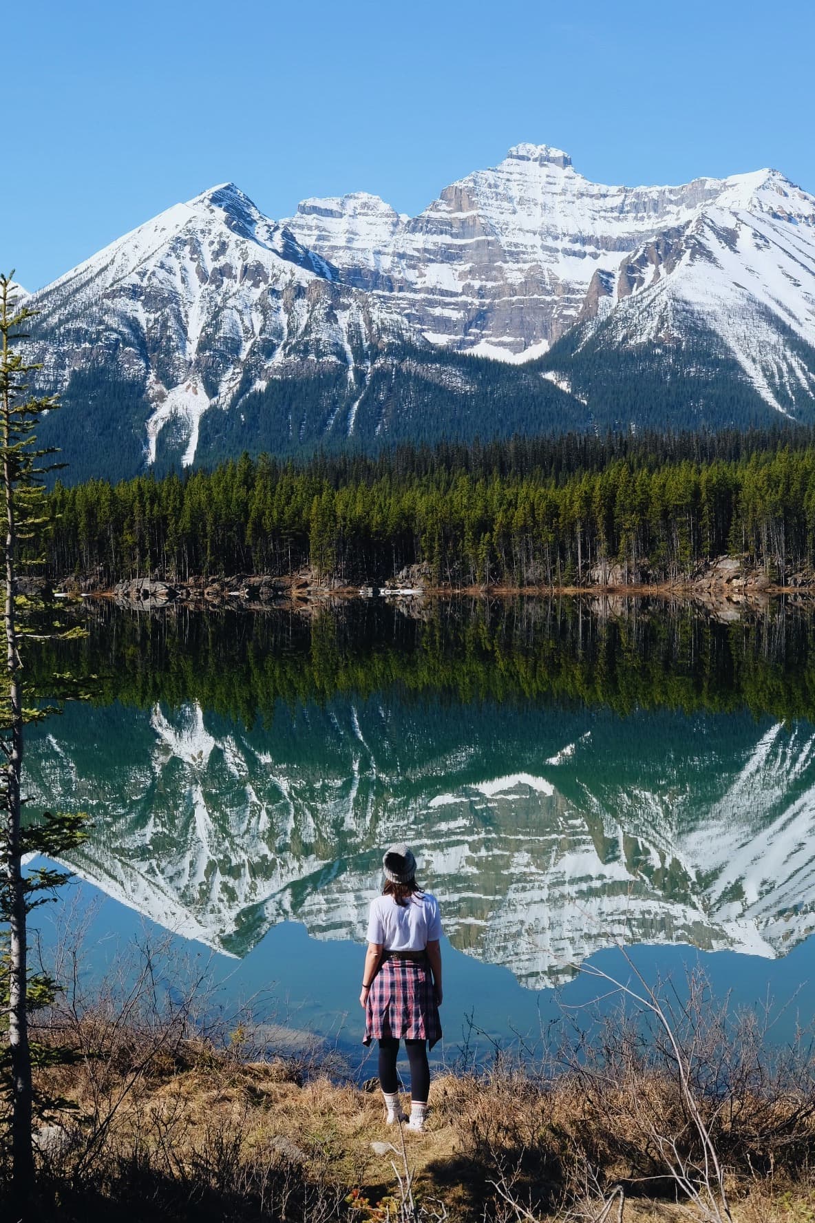 A woman standing in front of a clear lake with reflection of mountains in it.