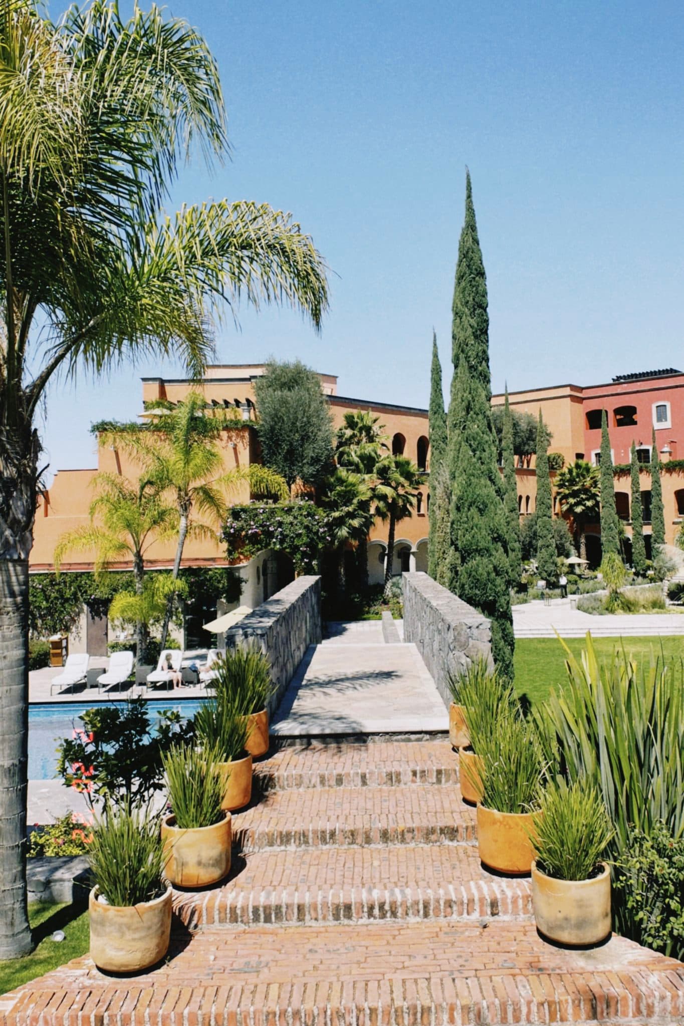 A view of a brick staircase lined with potted plants leading over a bridge to a large resort at Rosewood San Miguel de Allende during the day.