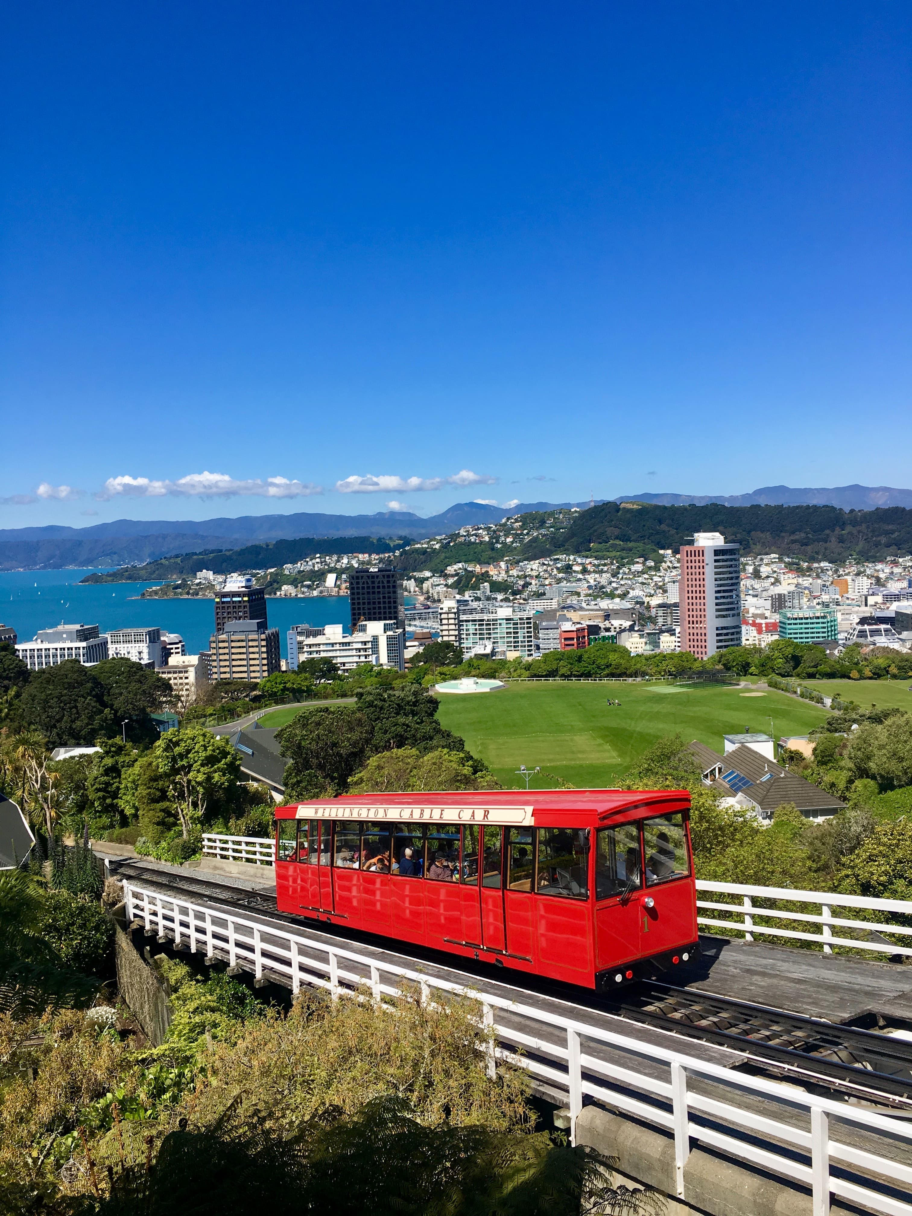 Red bus driving on a bridge with city buildings and sea in the background.