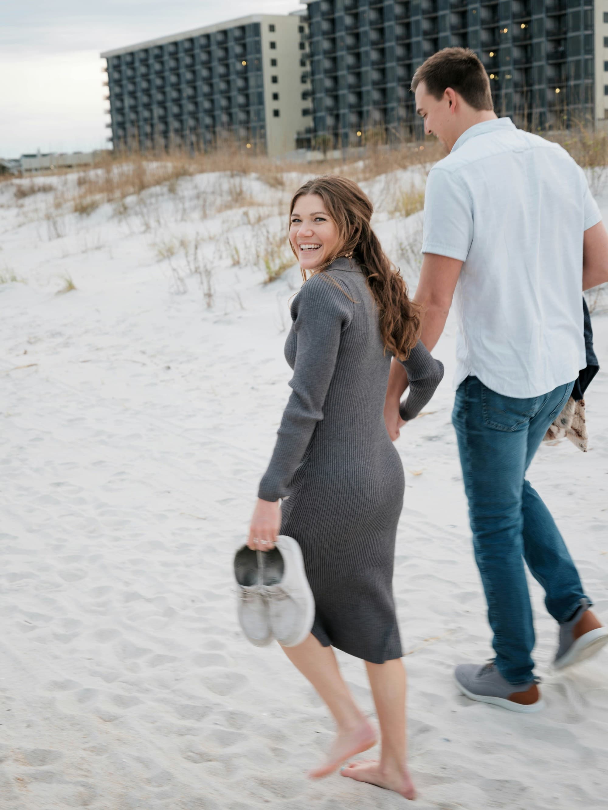The image shows two individuals walking on a sandy beach with buildings in the background,