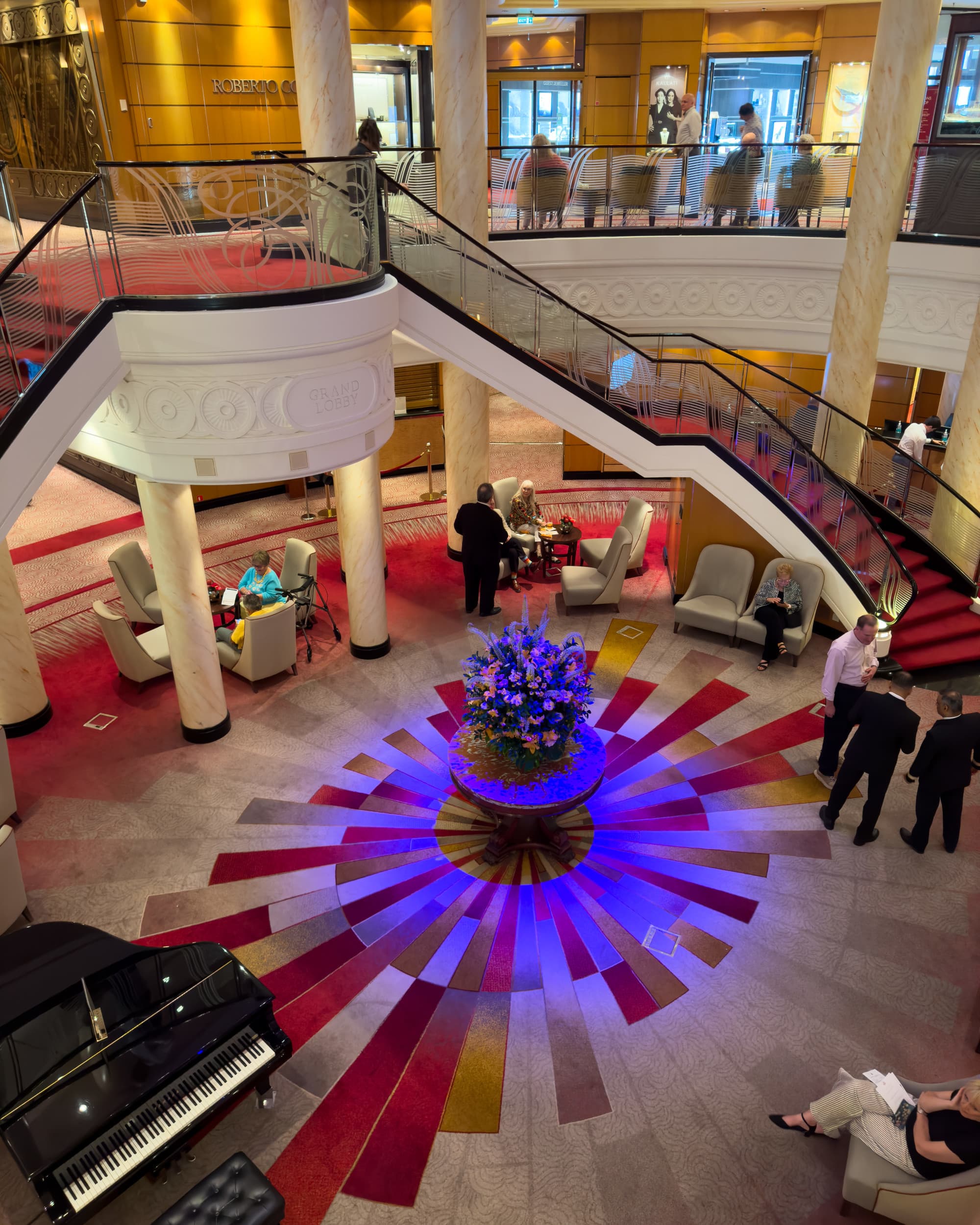 A view of the Grand Lobby from the second floor looking down, there are two curved black staircases and a circular red and blue pattern on the lower floor.