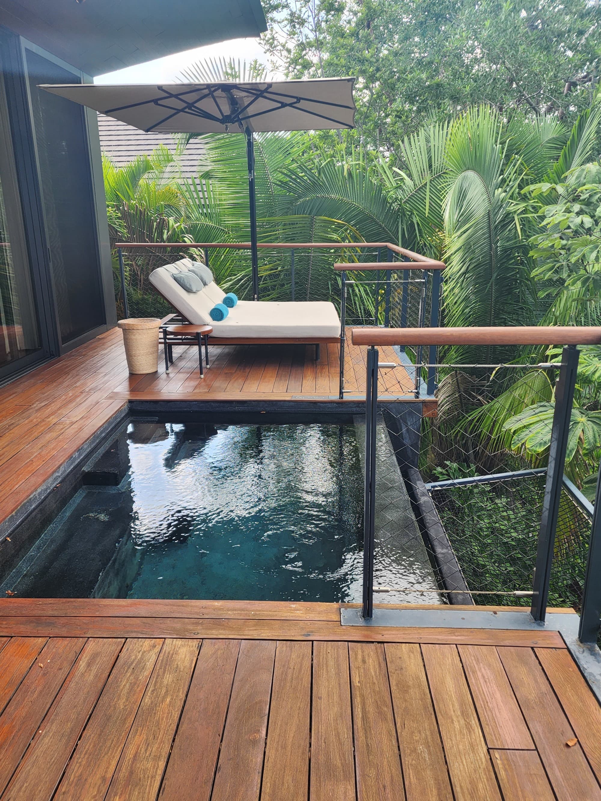 A view of a wooden deck with a small pool and a lounger with an umbrella with a lush forrest surrounding it.