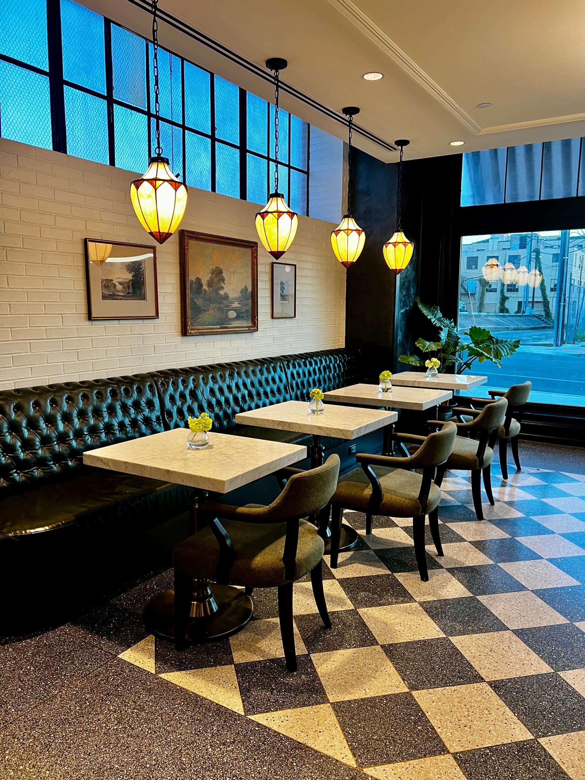A view of the dining area with black and white checkered floors, hanging lamps over each white table, black chairs and windows lining the top portion of the walls.