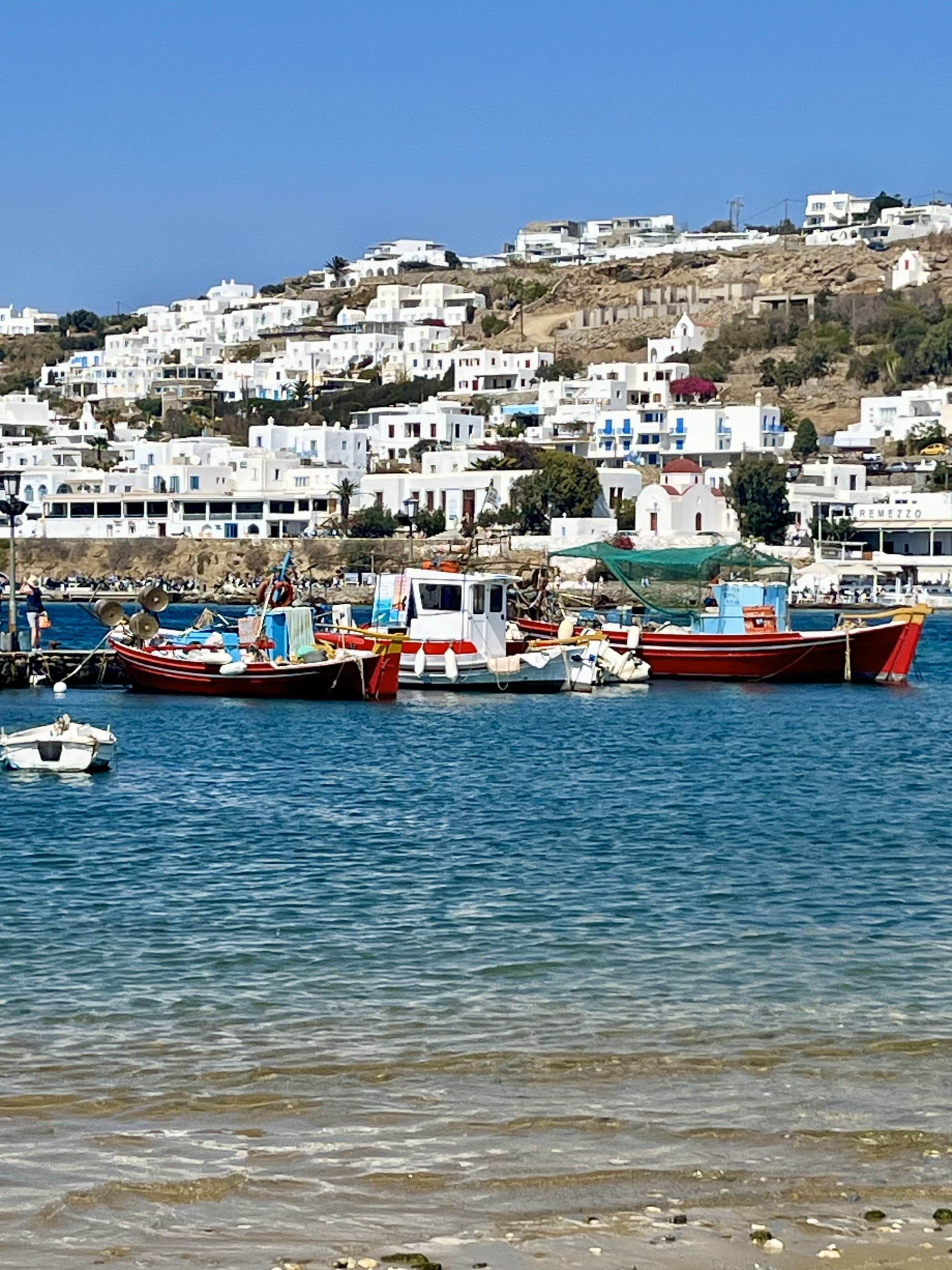 A view of boats in the ocean in Mykonos with white buildings lining the hill behind.