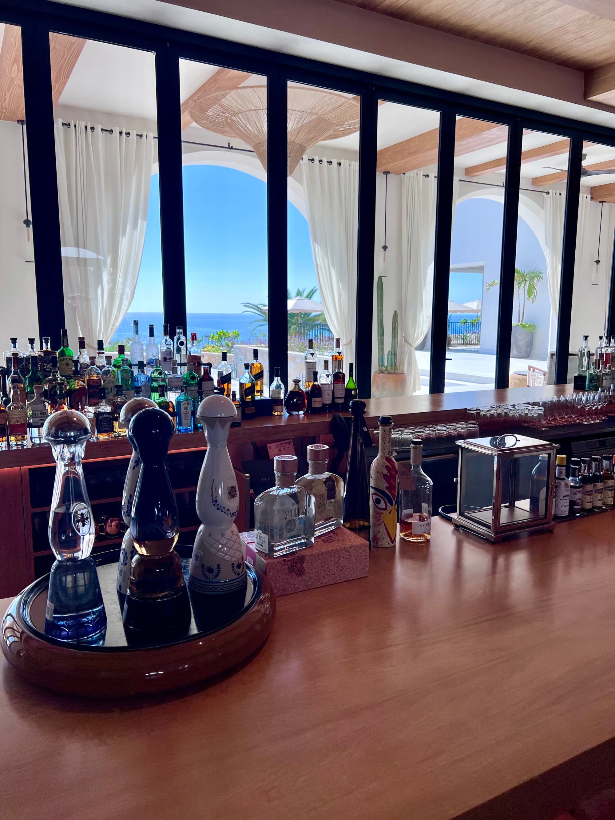 A close-up view of the bar La Casona with different bottles of tequila and other liquors, with large windows showing ocean views.