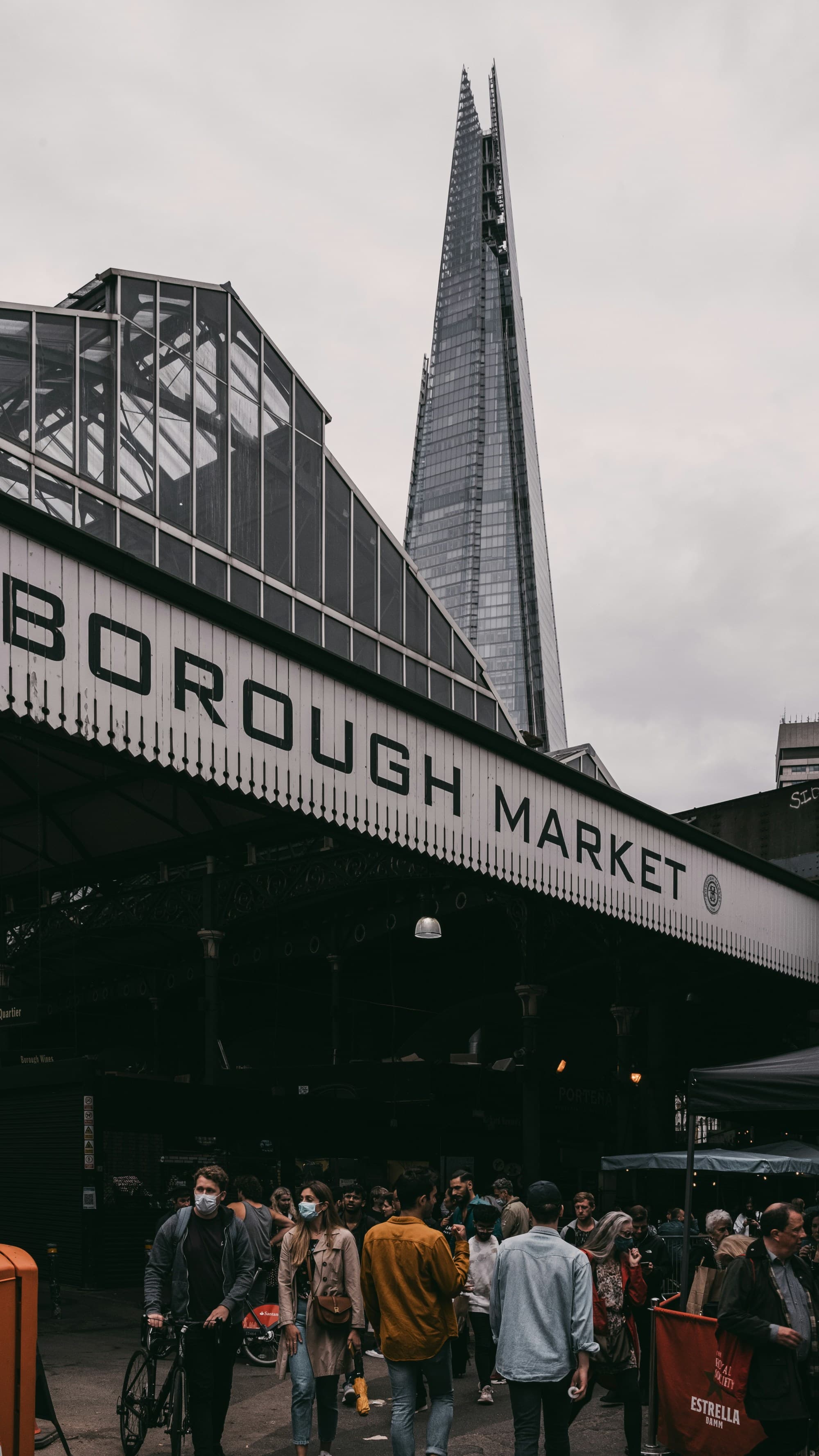 The image captures the vibrant essence of Borough Market, bustling with people and a cyclist under the iconic market sign, with the Shard looming in the background.