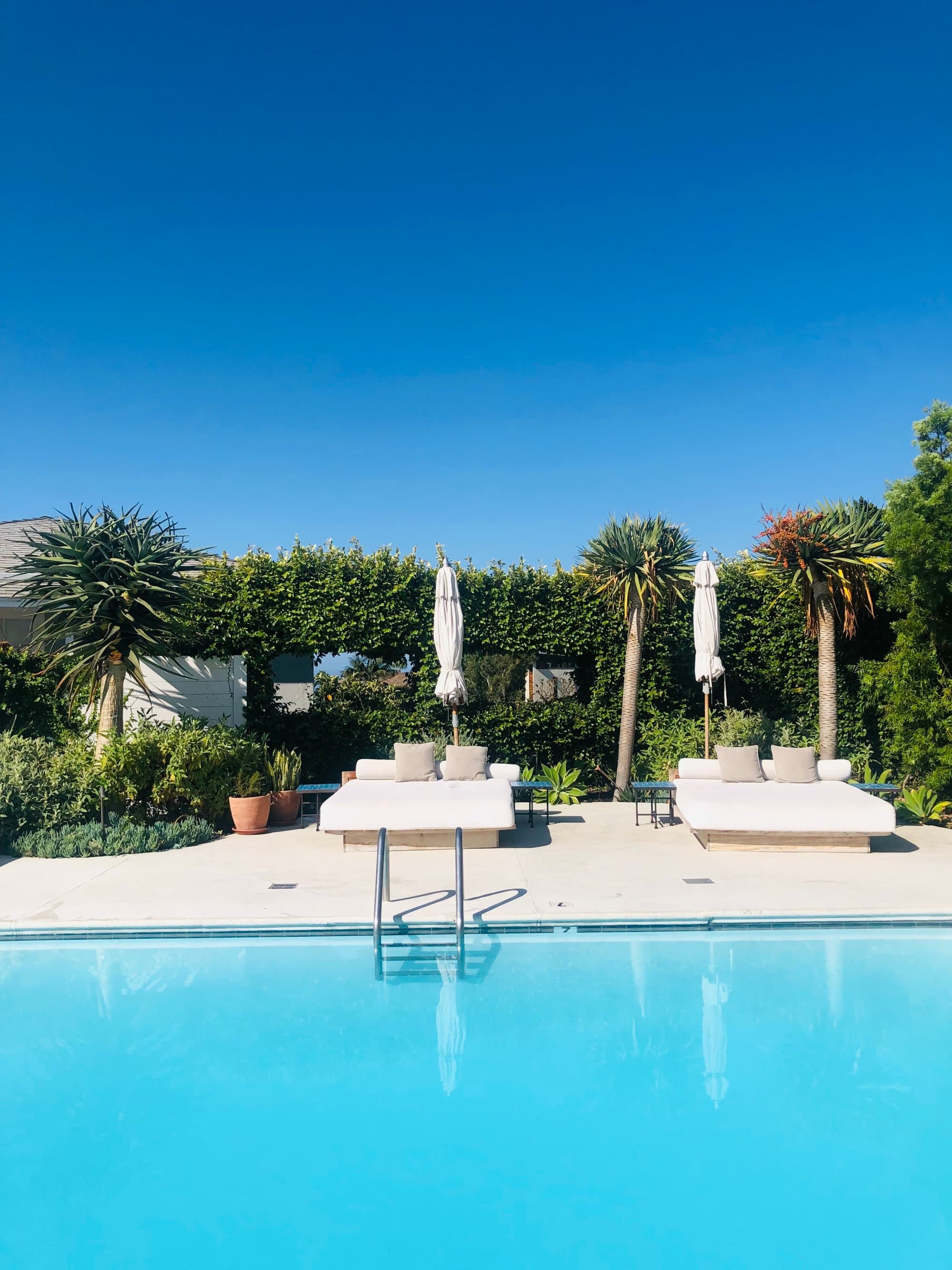An outdoor swimming pool with sun loungers and a neatly trimmed hedge under a clear blue sky.