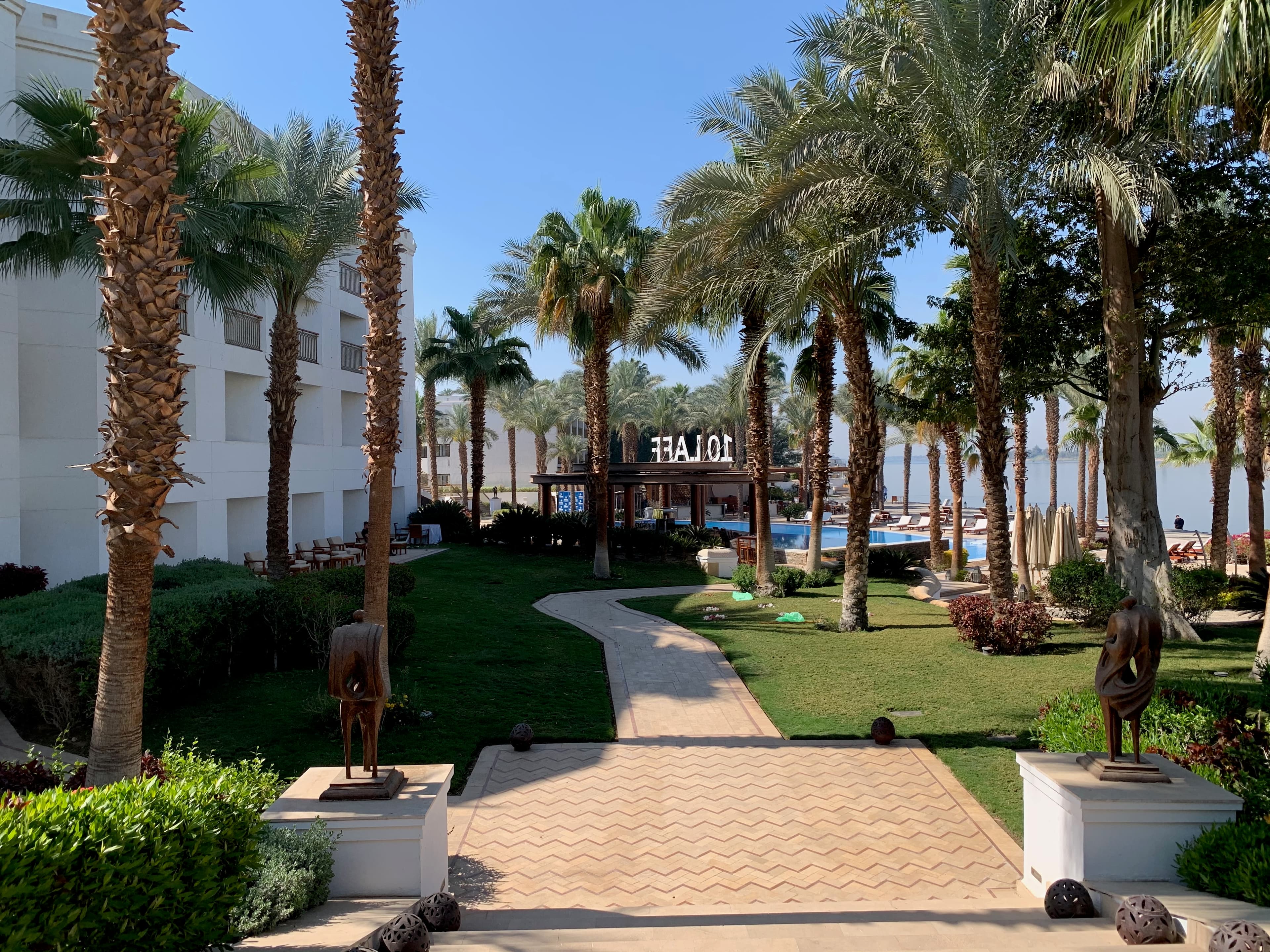 The image presents a landscaped pathway lined with palm trees and sculptures, leading to a building.