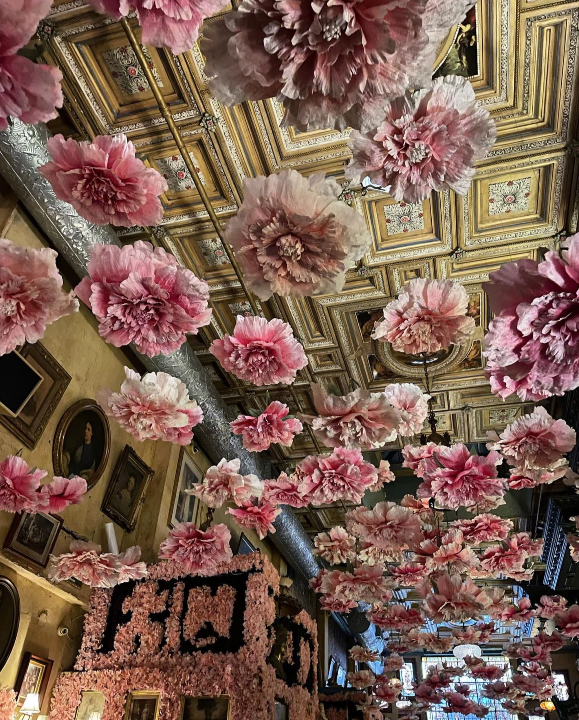 An elegant ceiling with golden patterns and walls adorned with art, contrasted by numerous pink flowers hanging upside down.