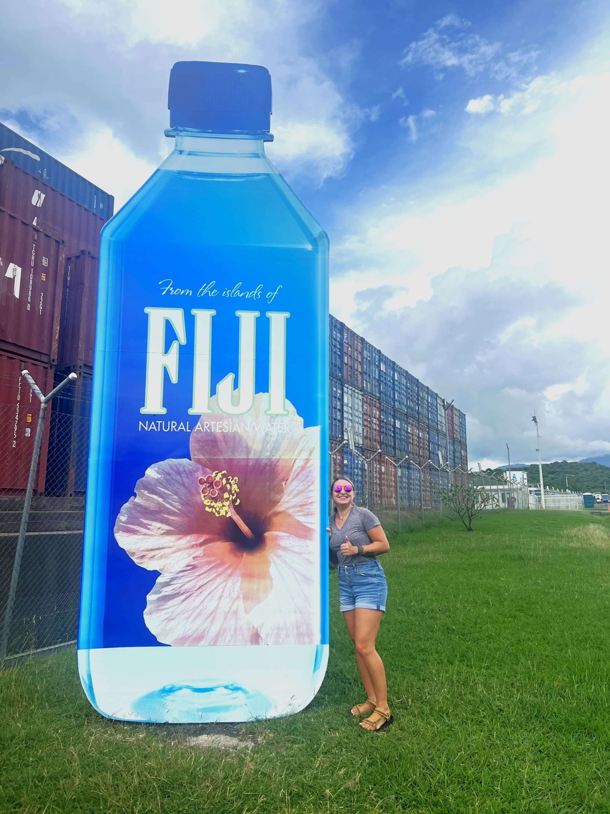 Danielle standing next to a large advertisement of a Fiji water bottle, with a clear sky and containers in the background.