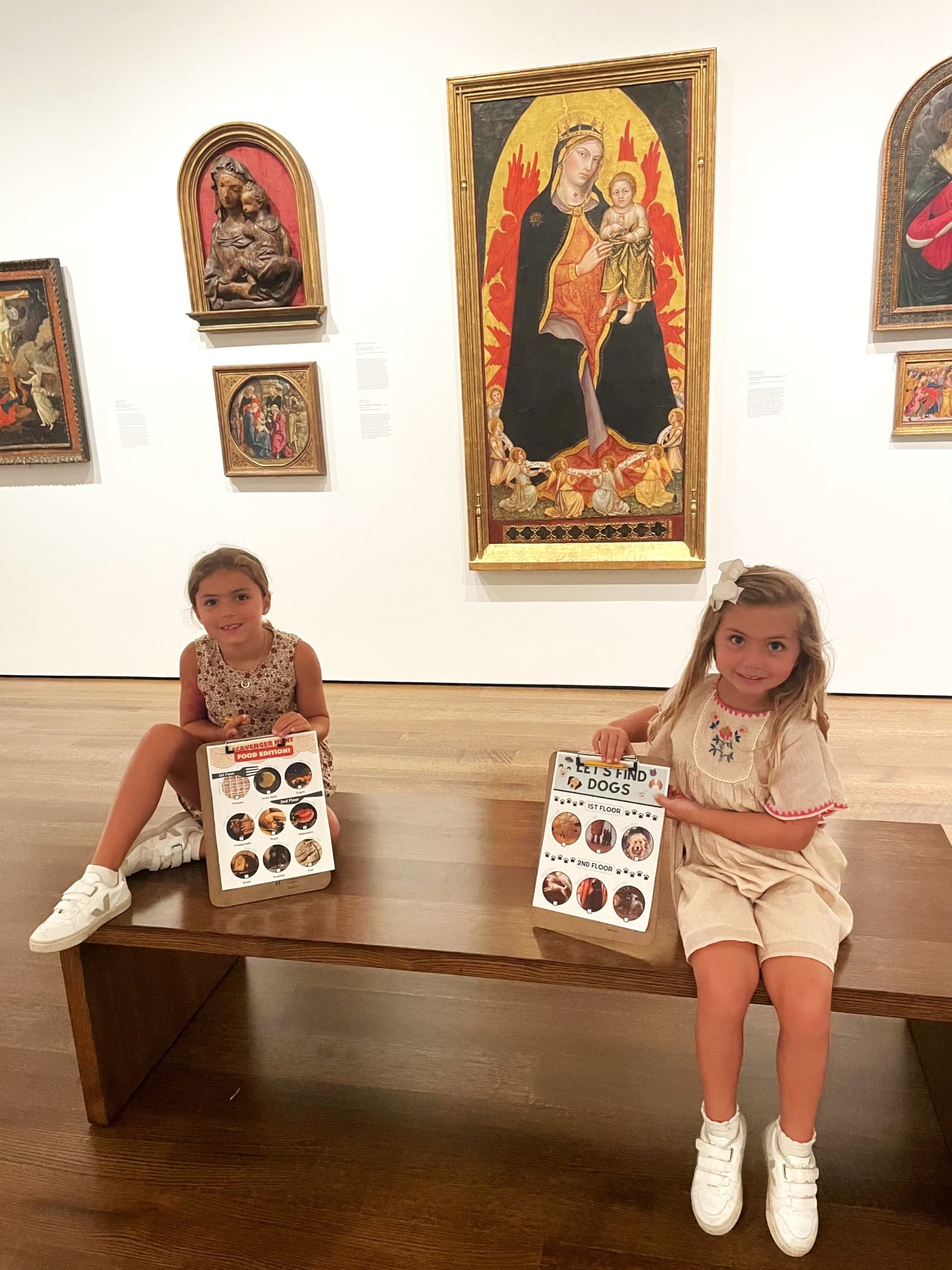 Two kids sitting on a bench in an art museum, holding "Let's Find Dogs" scavenger hunt clipboards.