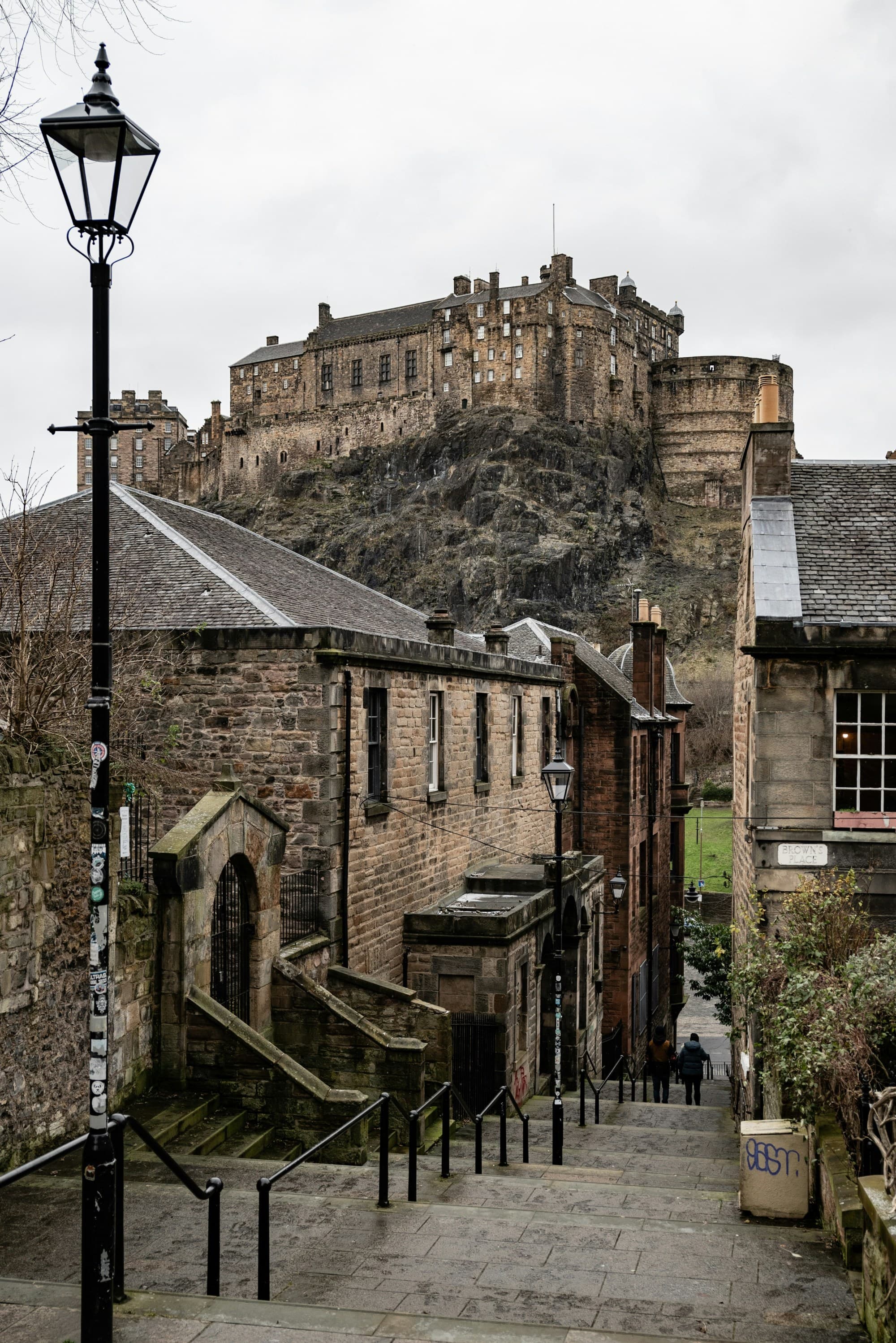 The image presents a picturesque cobbled street leading to an imposing castle atop a hill, with an old street lamp standing guard under overcast skies.