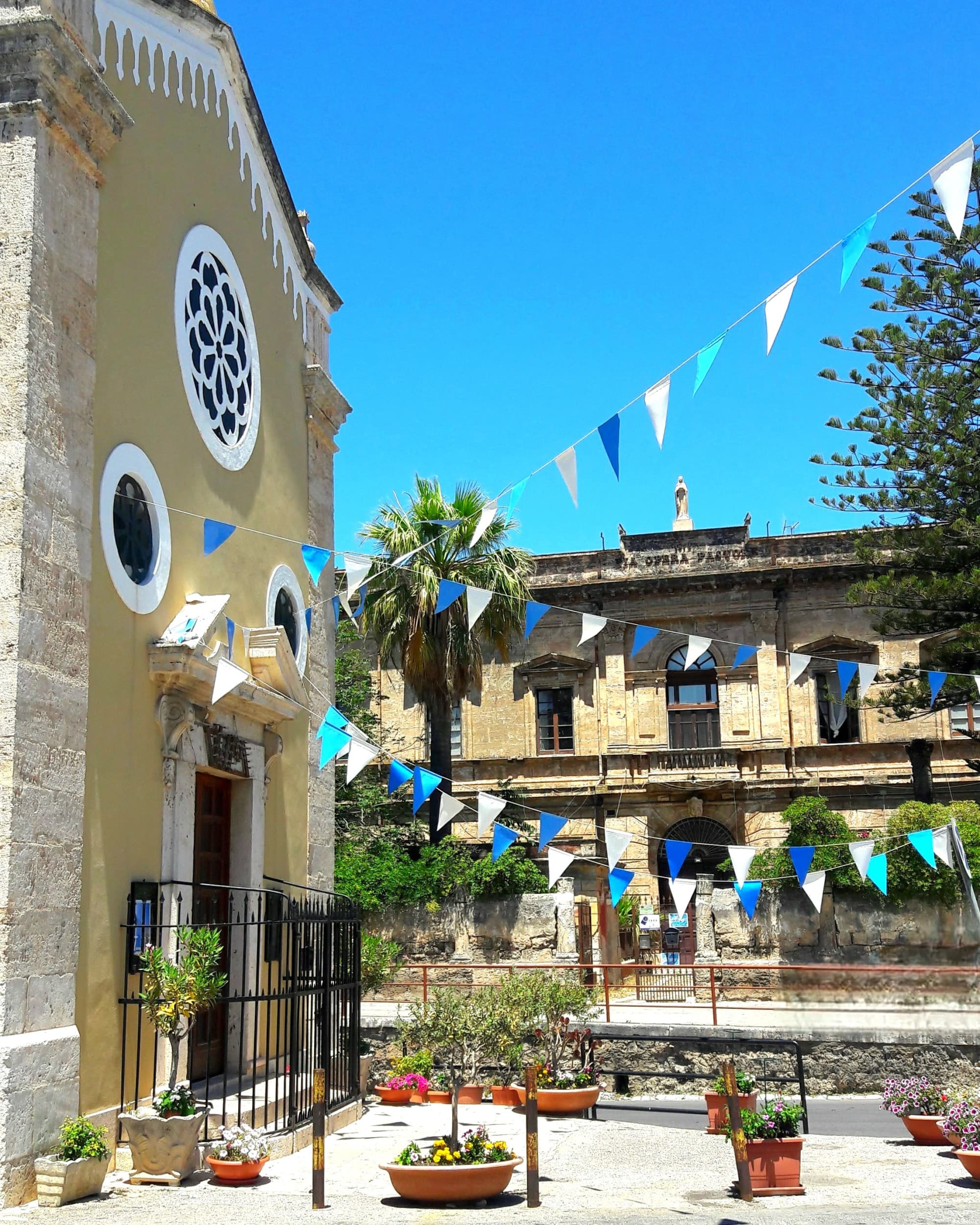 A concrete walkway lined with plants leading to a yellow building, with blue and yellow flags flying outside, which you might see on a Sicily itinerary, 10 days. - Ali Lefebvre