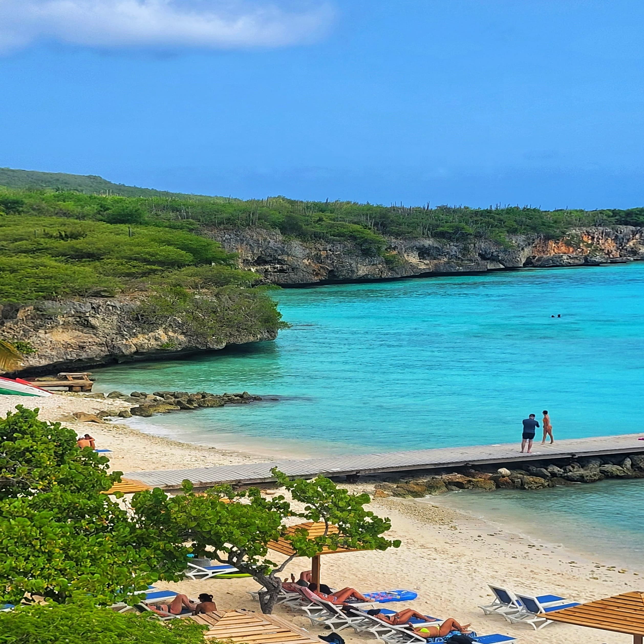 A serene tropical beach with a pier going out onto clear turquoise waters, lush greenery and lounge chairs on the sand.