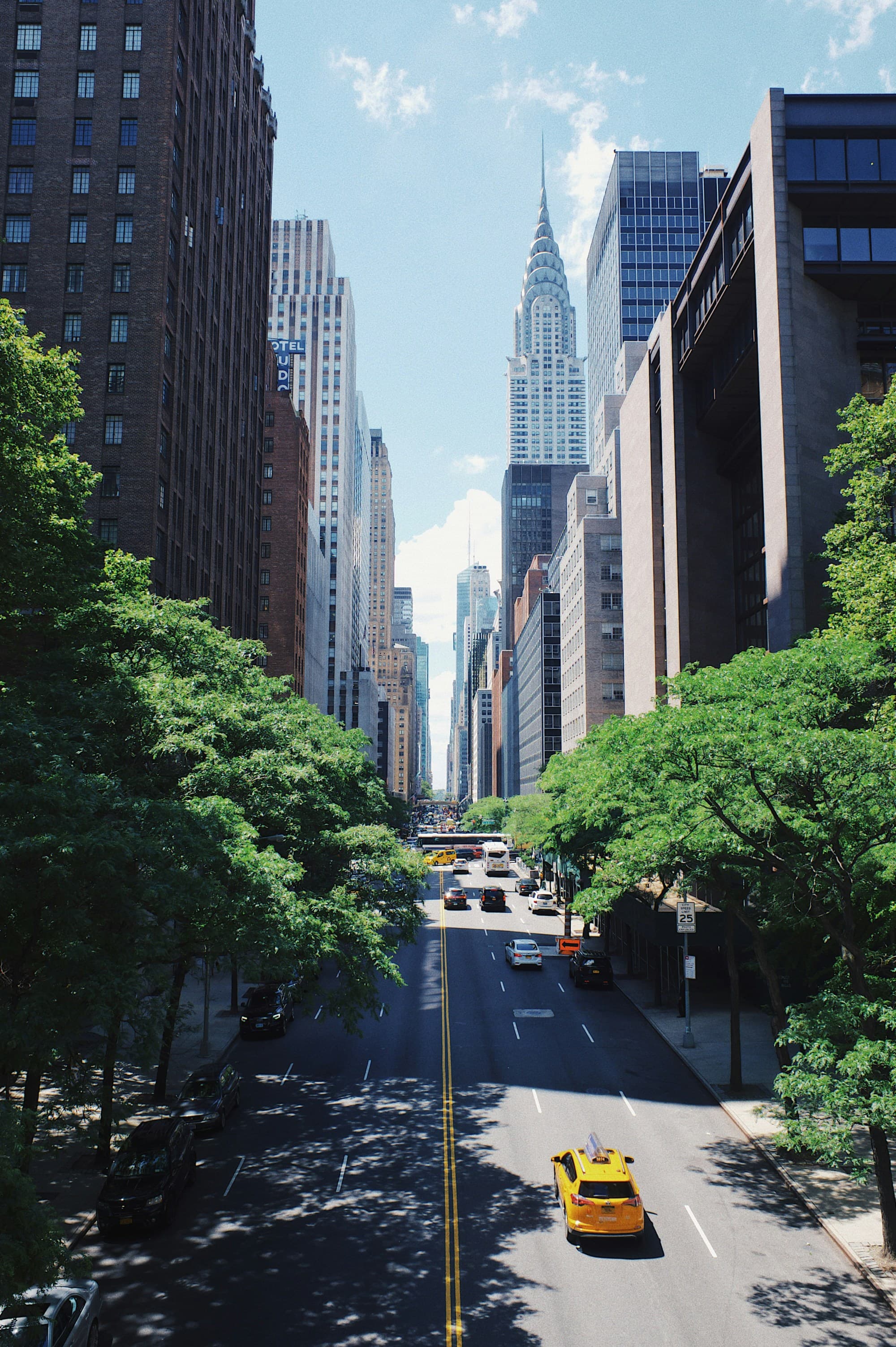 A bustling New York City street with greenery and the Chrysler Building in the distance under a clear blue sky.