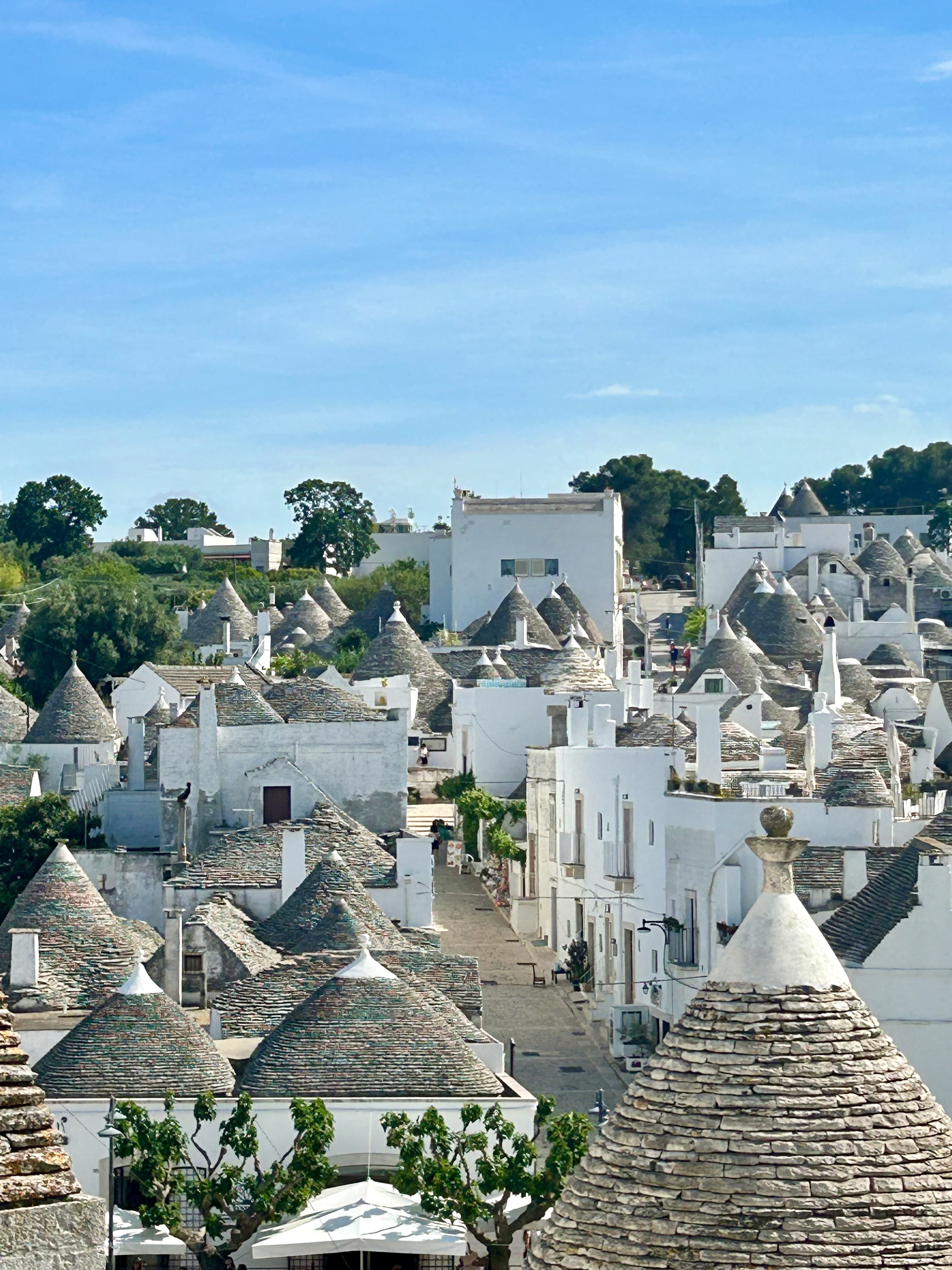 The image offers a scenic view of Alberobello, showcasing its distinctive trulli structures under a clear sky.