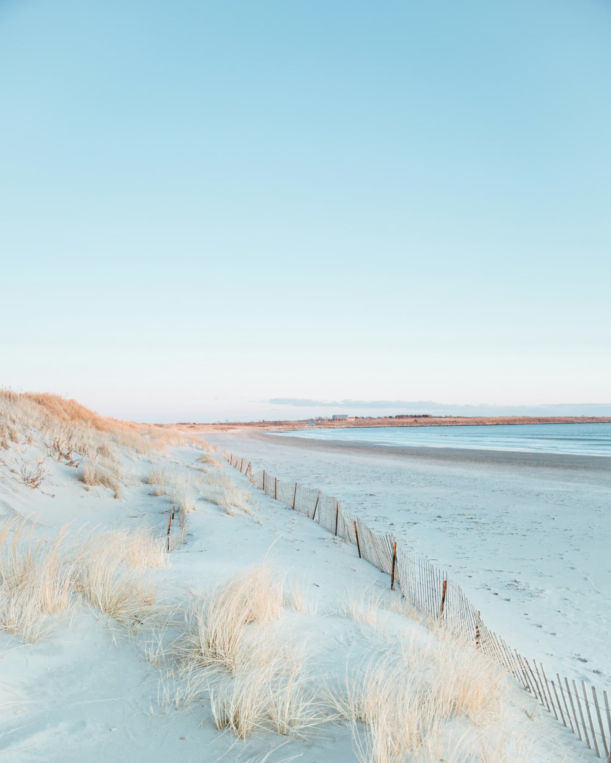 A serene beach scene, with sand dunes and sparse vegetation framed by a clear sky and a rustic wooden fence.