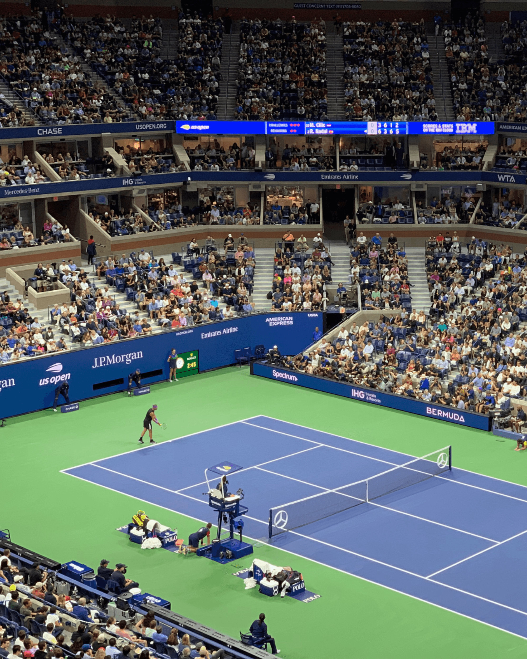 A tennis match with spectators, featuring a blue court, players in action, and professional tournament scoreboards.