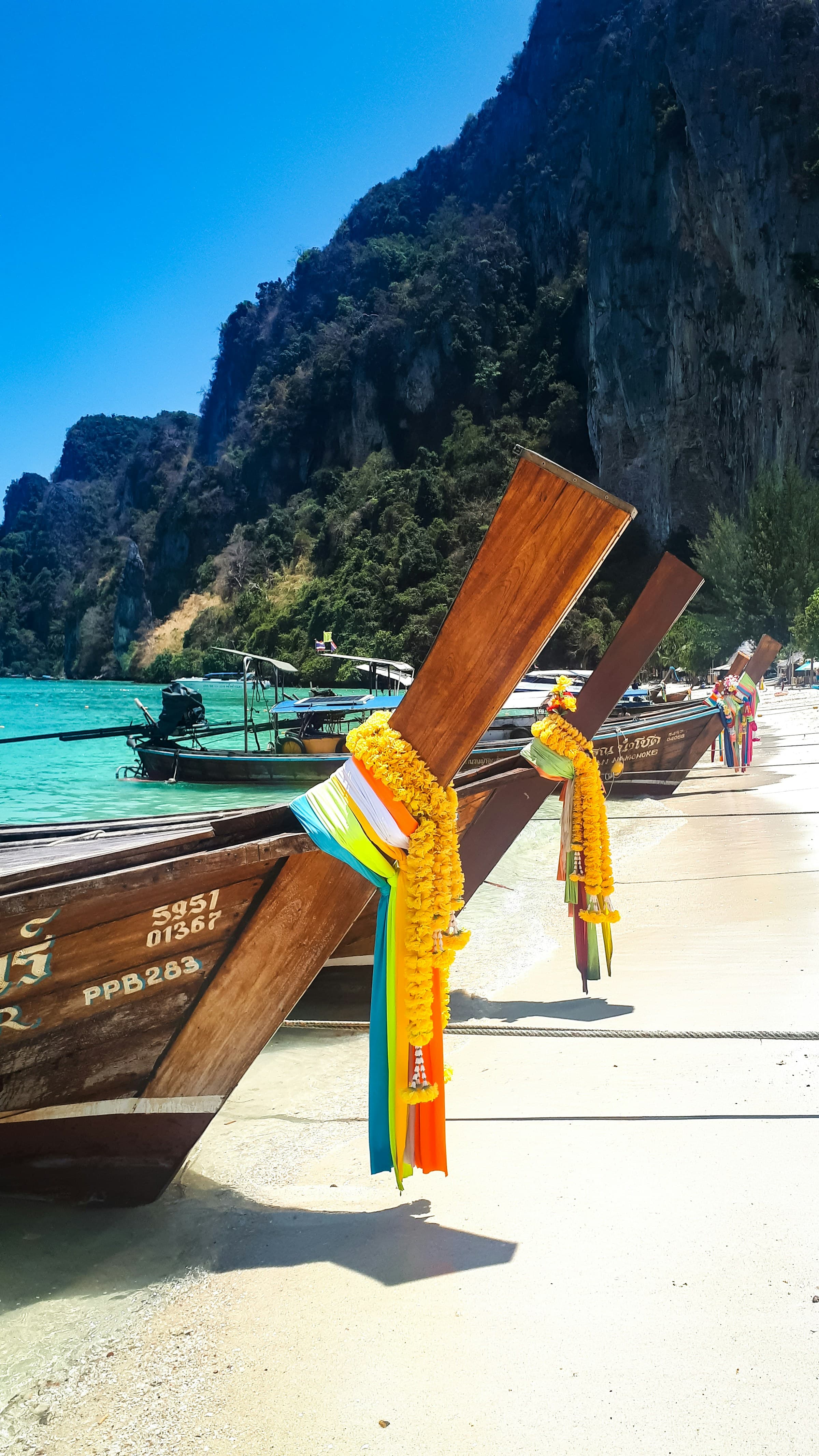 This image depicts wooden boats lined up on a white sandy beach with turquoise blue water and large mountains in the background.