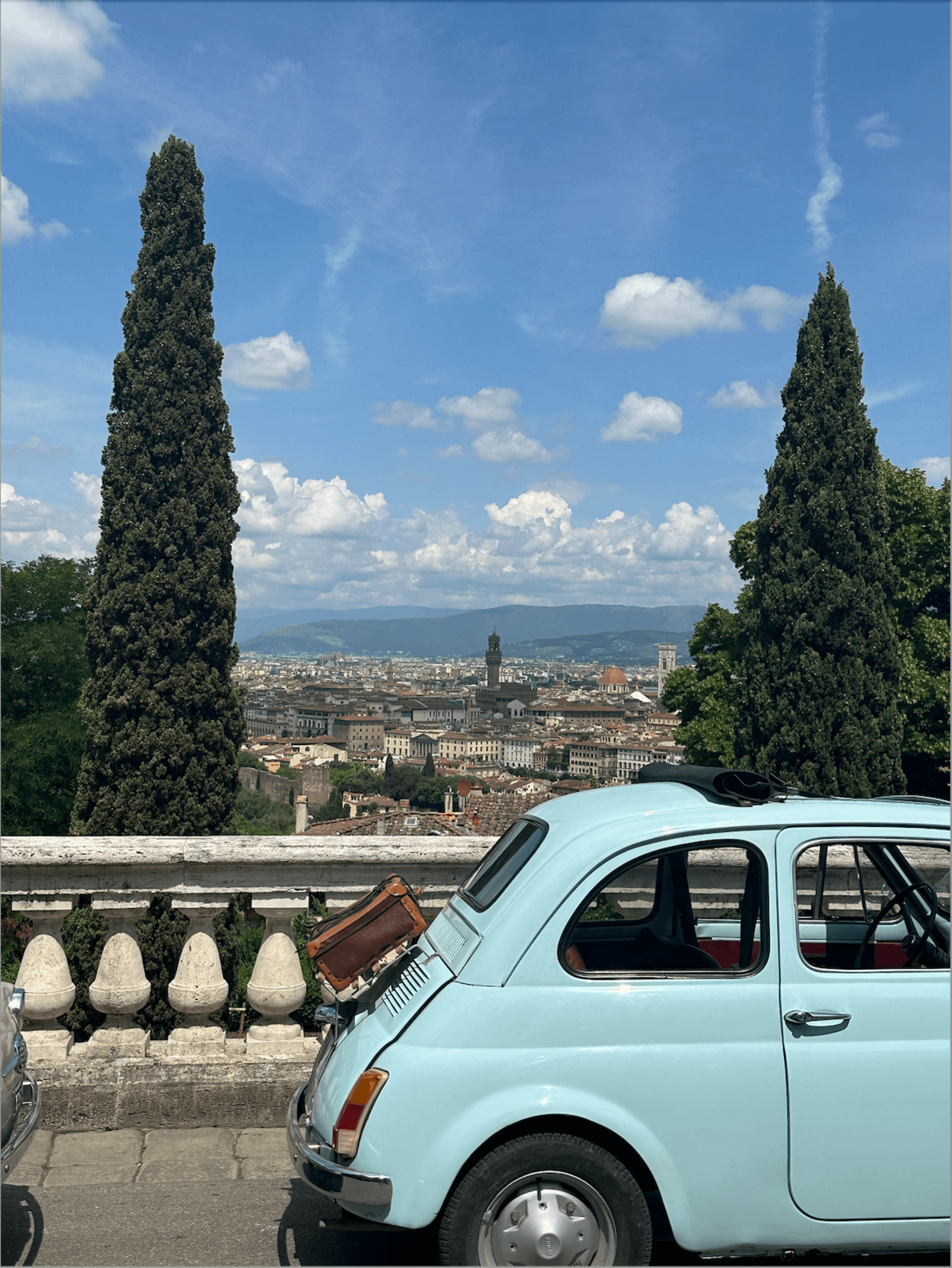 A vintage blue car adds a touch of nostalgia to a scenic overlook of a tree-lined cityscape under cloudy skies.