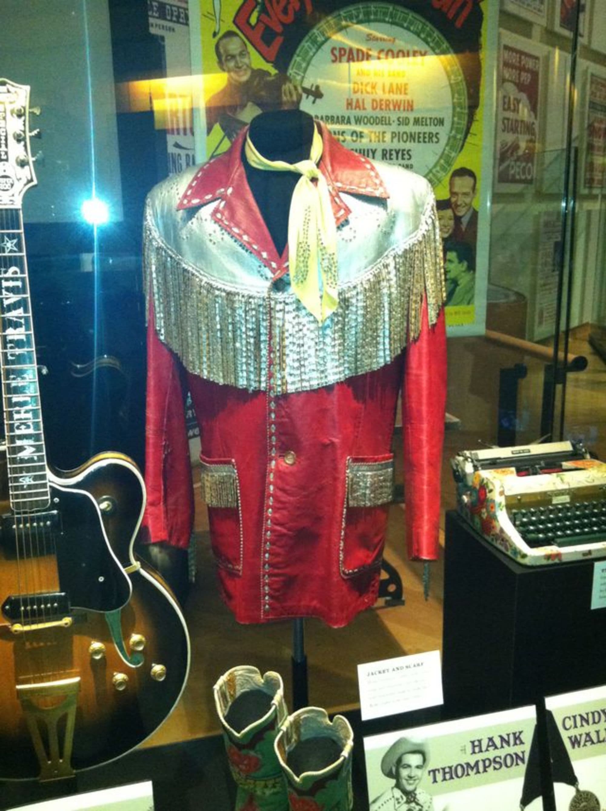 A red, vintage stage costume with silver details, alongside a guitar and memorabilia in a display case