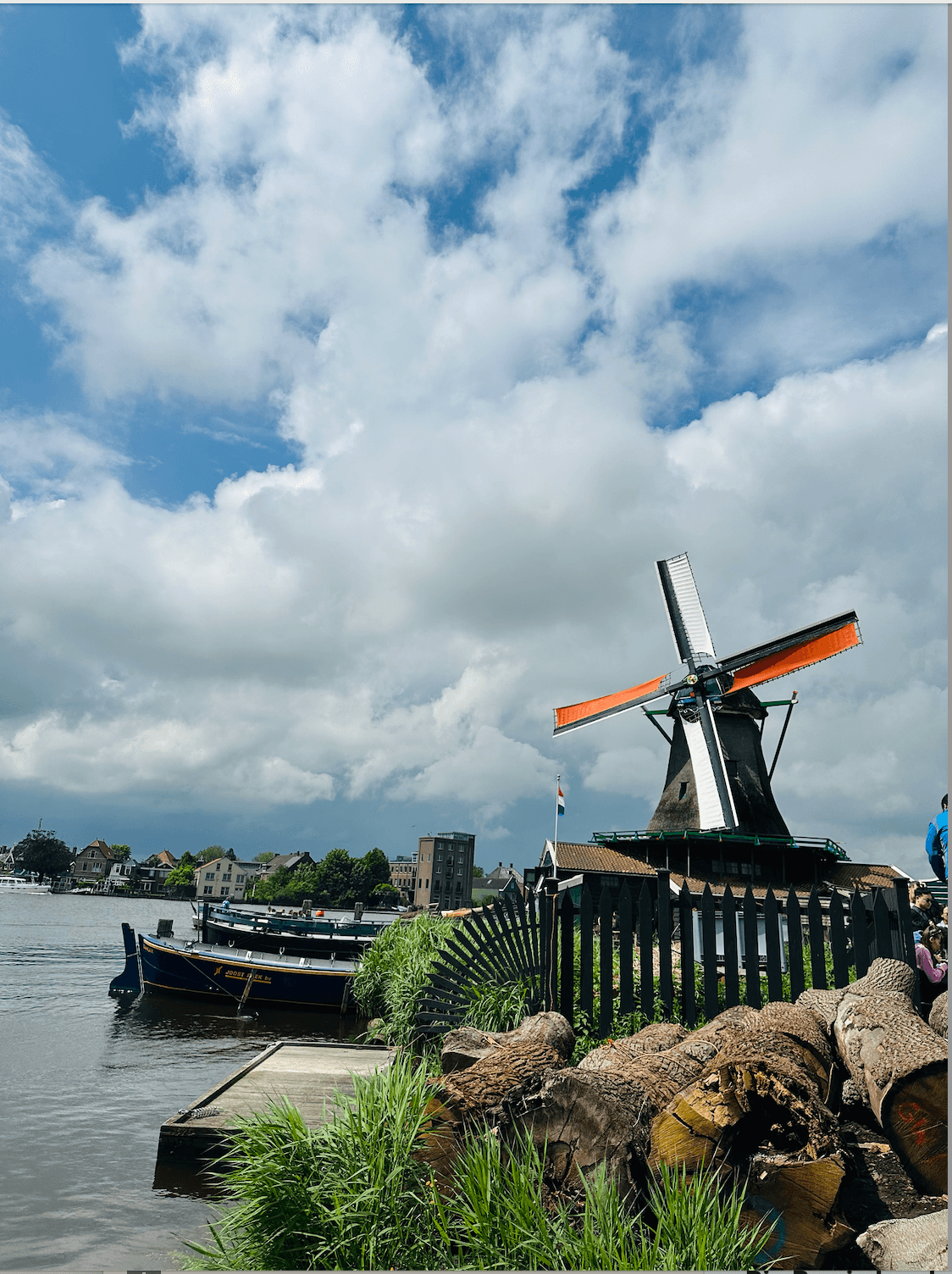 A windmill standing on the edge of a body of water with canoes parked next to it and buildings in the distance along the shore line.