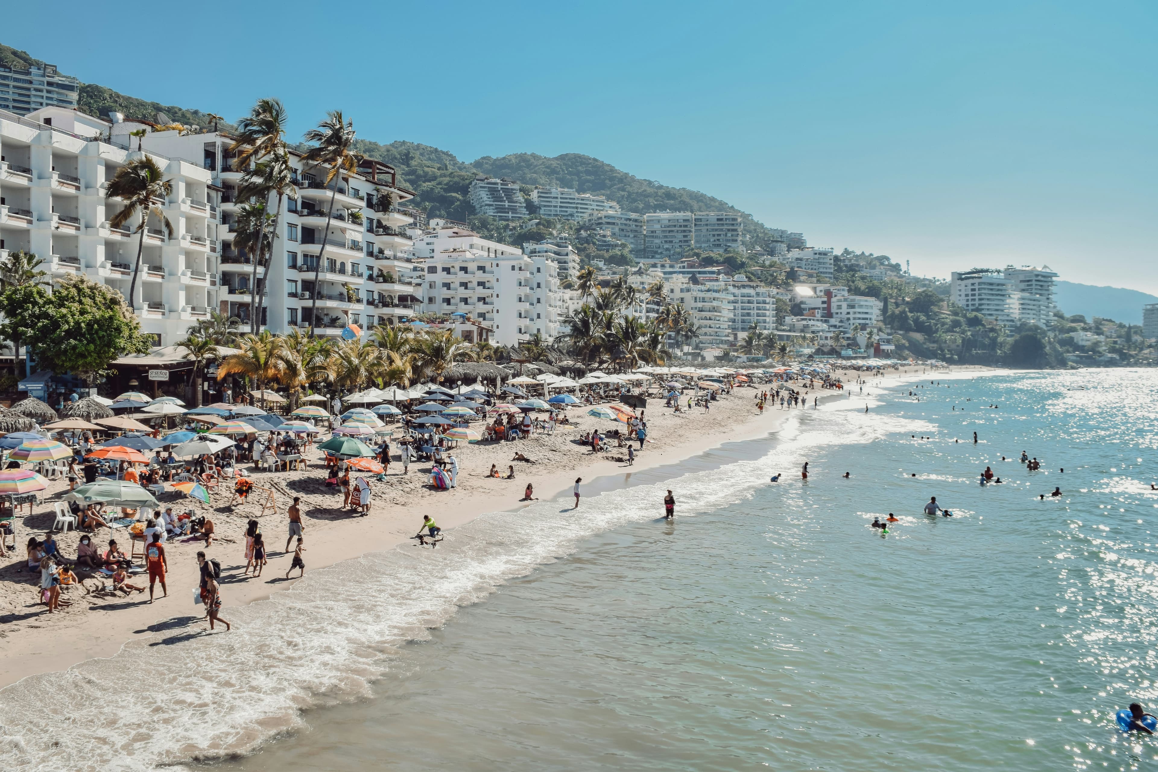 A view of a busy beach with people in the ocean and buildings and mountains behind