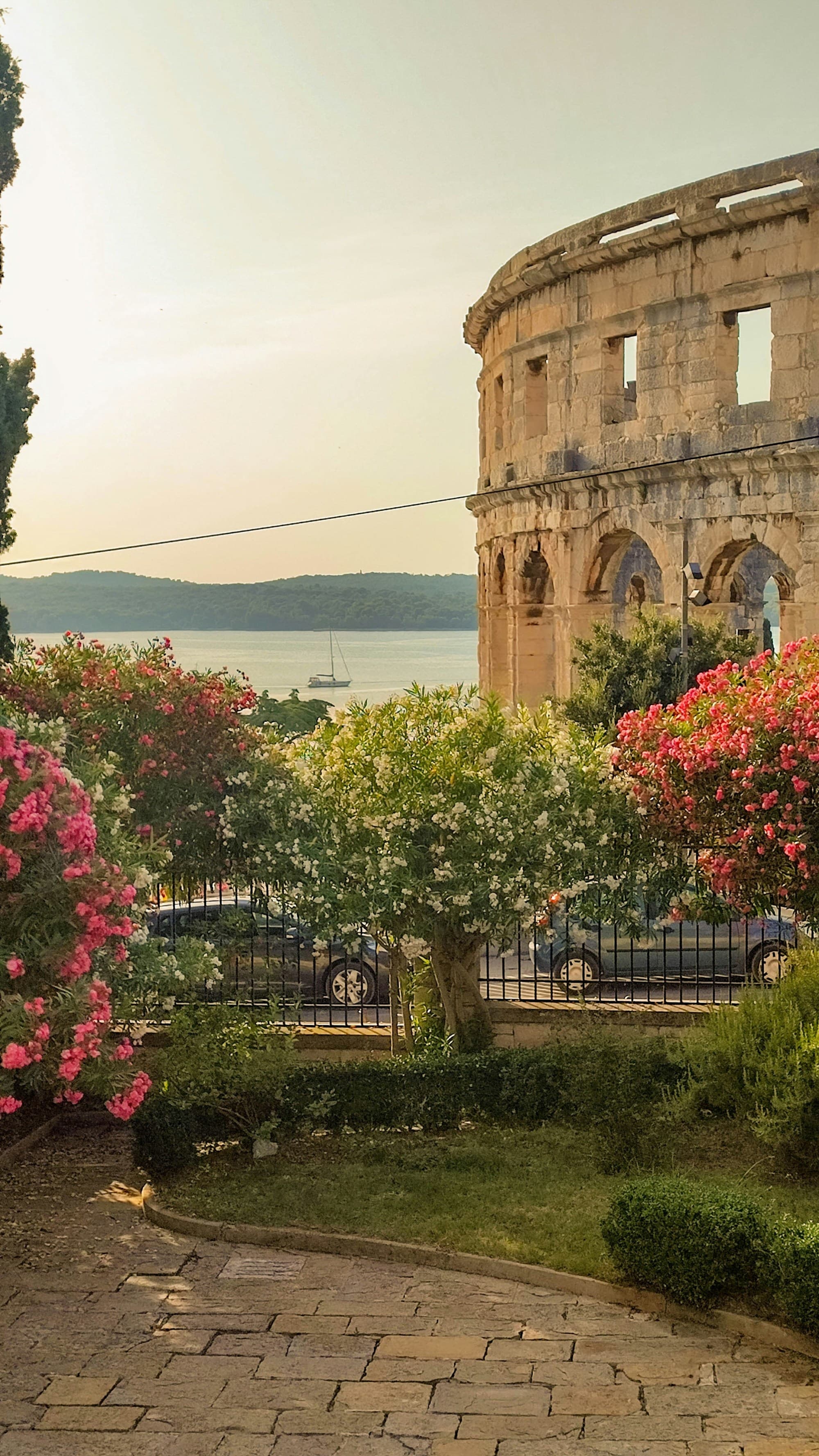 A side view of the exterior of the Roman amphitheater in Pula, with flowers and bushes in the forefront, and sea and mountains in the background.