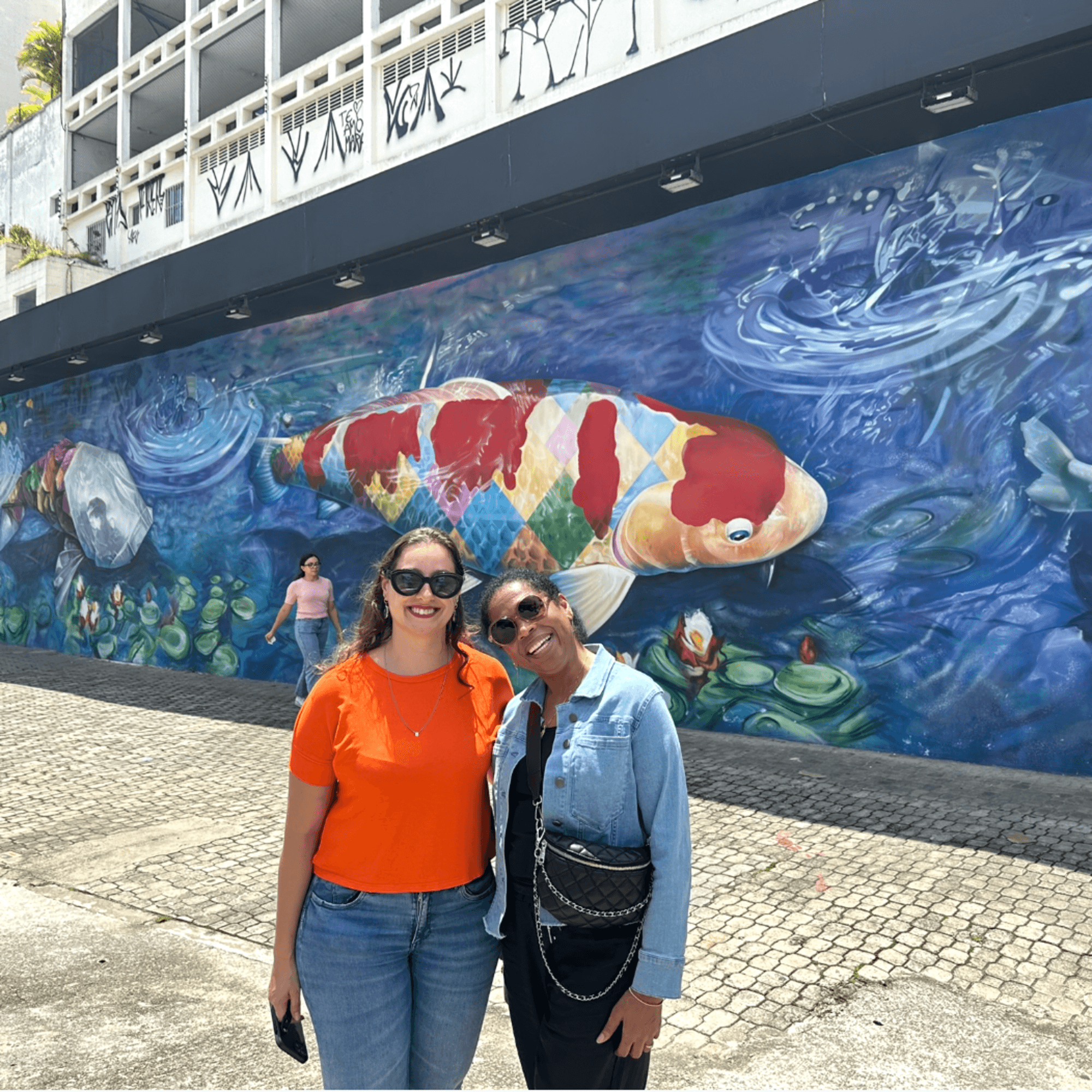 The image captures two people posing before a colorful mural depicting a large koi fish surrounded by marine life.