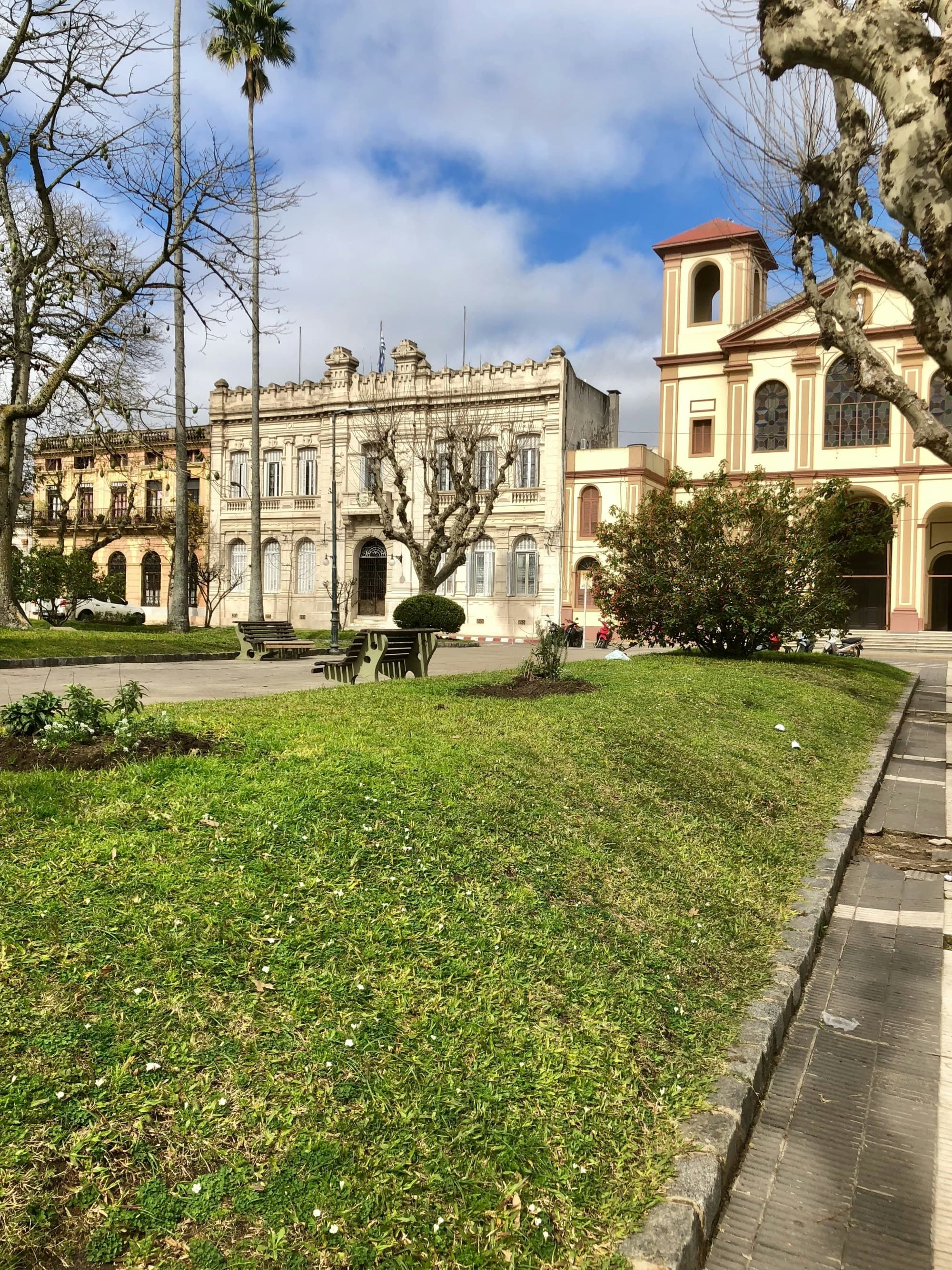 A classical building with a green lawn, adjacent to a smaller religious structure, under a partly cloudy sky.