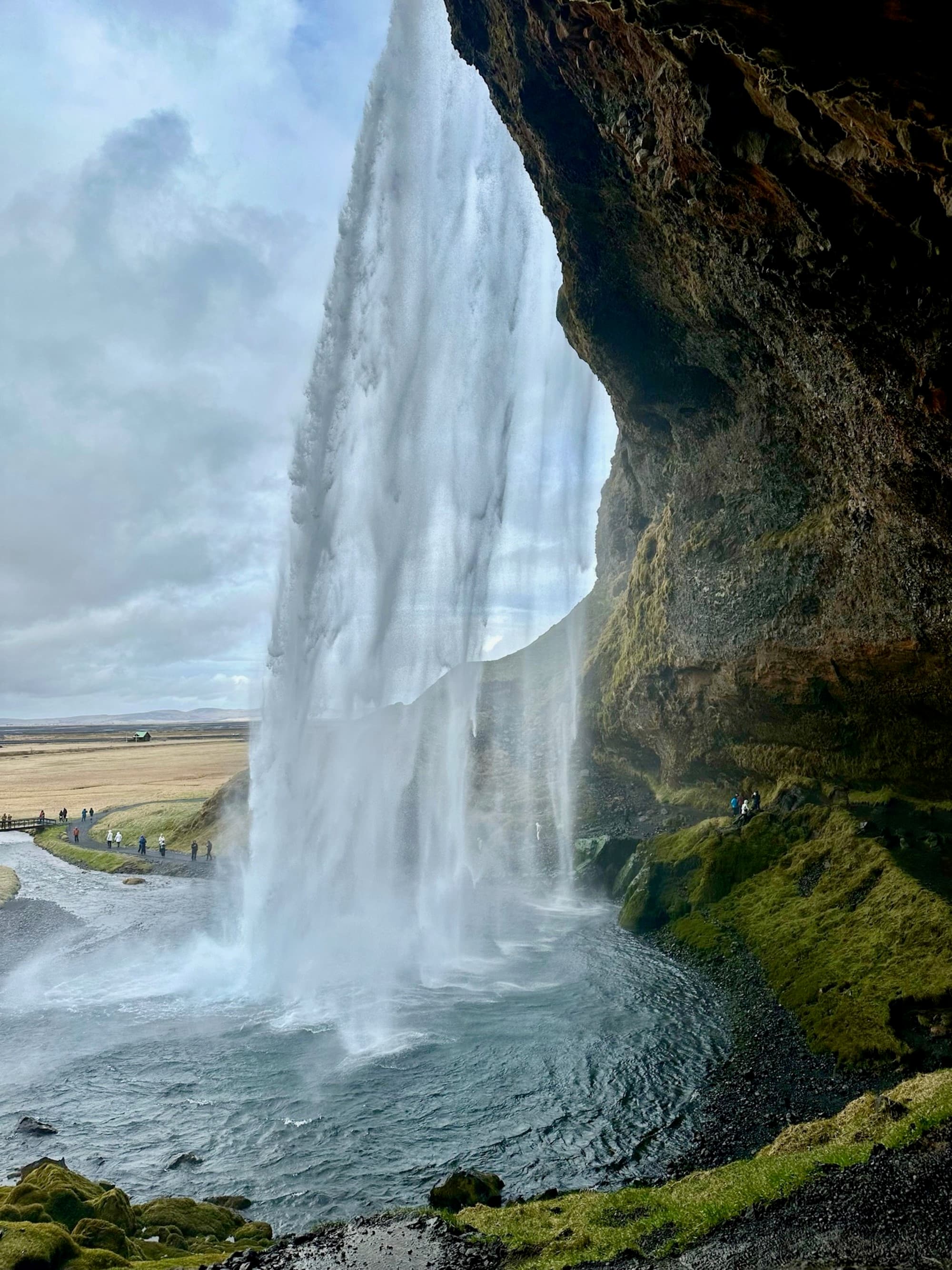 A majestic waterfall cascades down a cliff with a cave visible behind the water curtain, amidst lush greenery.