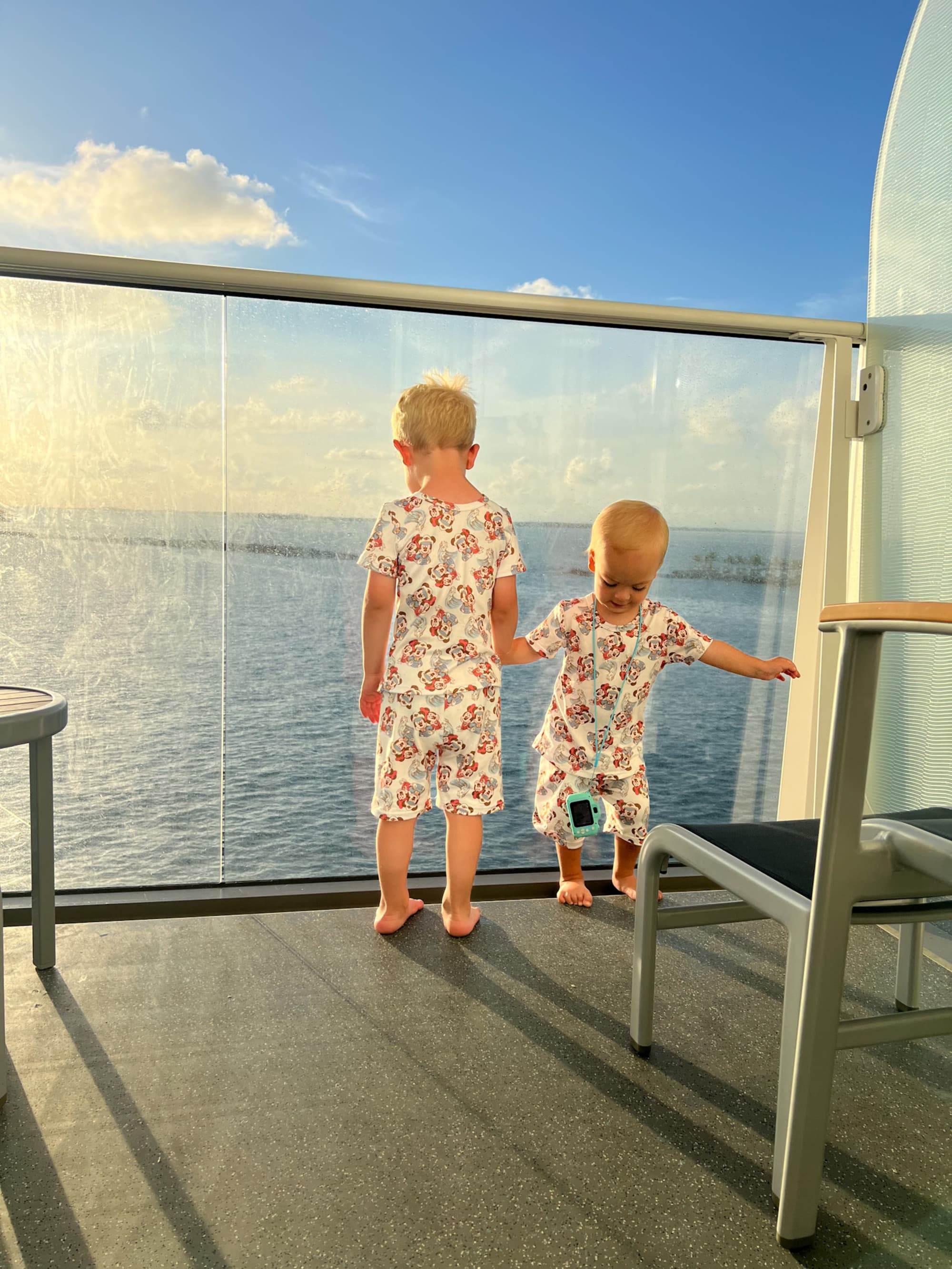 The image shows two kids in matching outfits, enjoying a sunny day by the sea behind a glass barrier on a cruise.