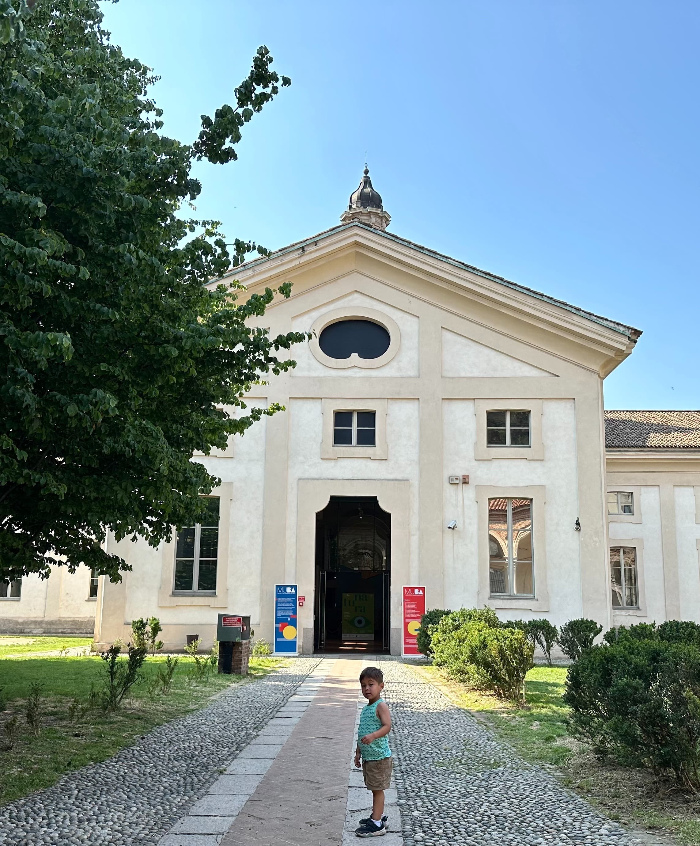 A child standing on a tree-lined path in front of a white building in the MUBA (Museo dei Bambini Milano) Gardens