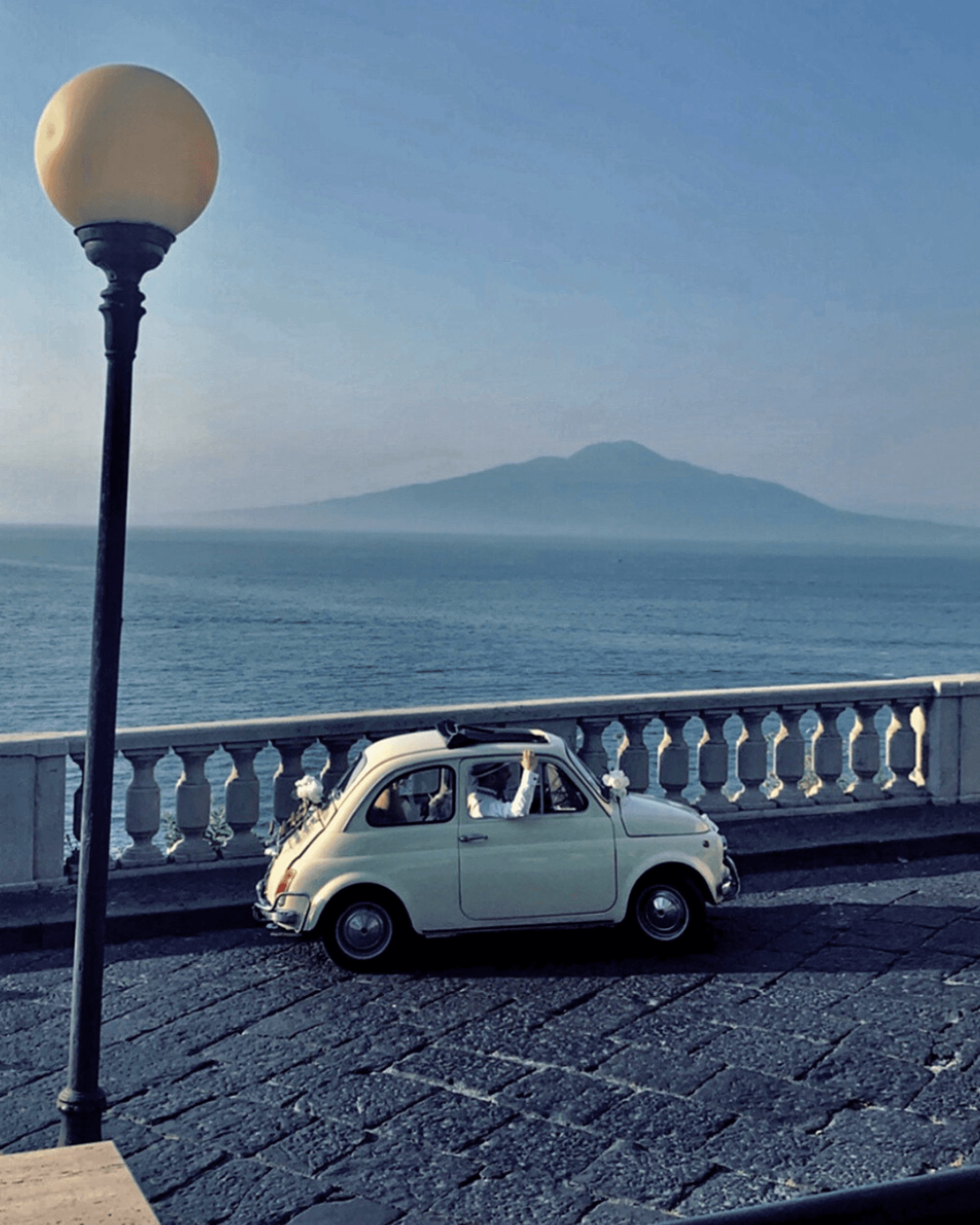 The image features a classic car parked by the seaside, with a street lamp and a large mountain in the background.