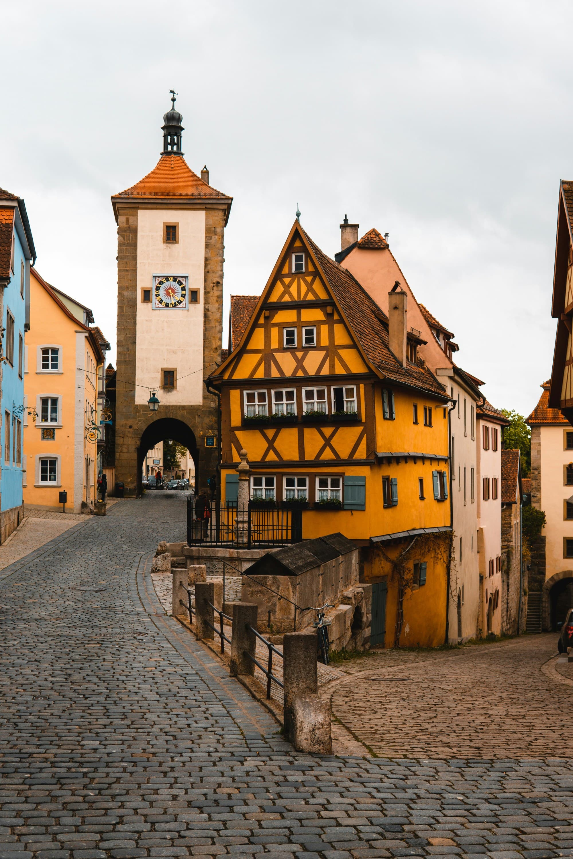 The image captures a charming cobblestone street leading to an old clock tower, lined with colorful traditional European buildings.